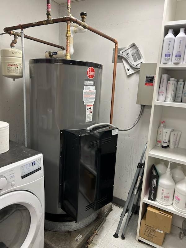 Water heater and laundry appliances in a utility room. Copper pipes, metal tank, white walls.