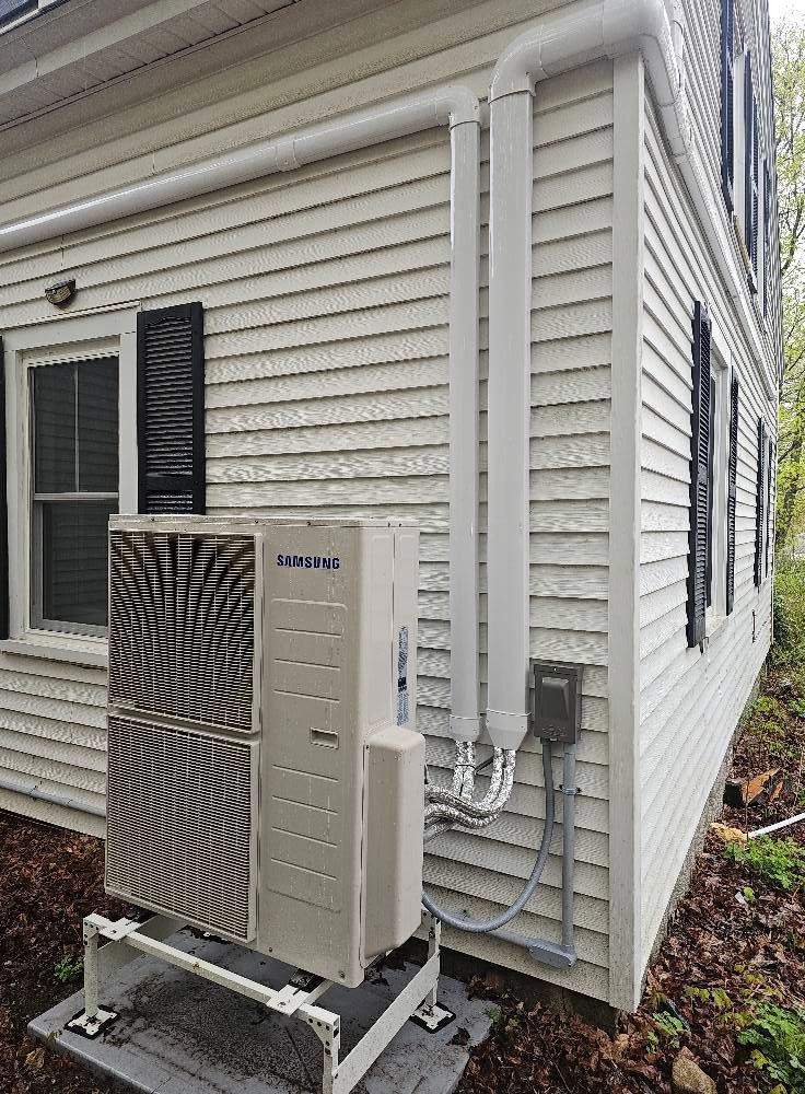 An outdoor HVAC unit on a white house, with vent pipes.