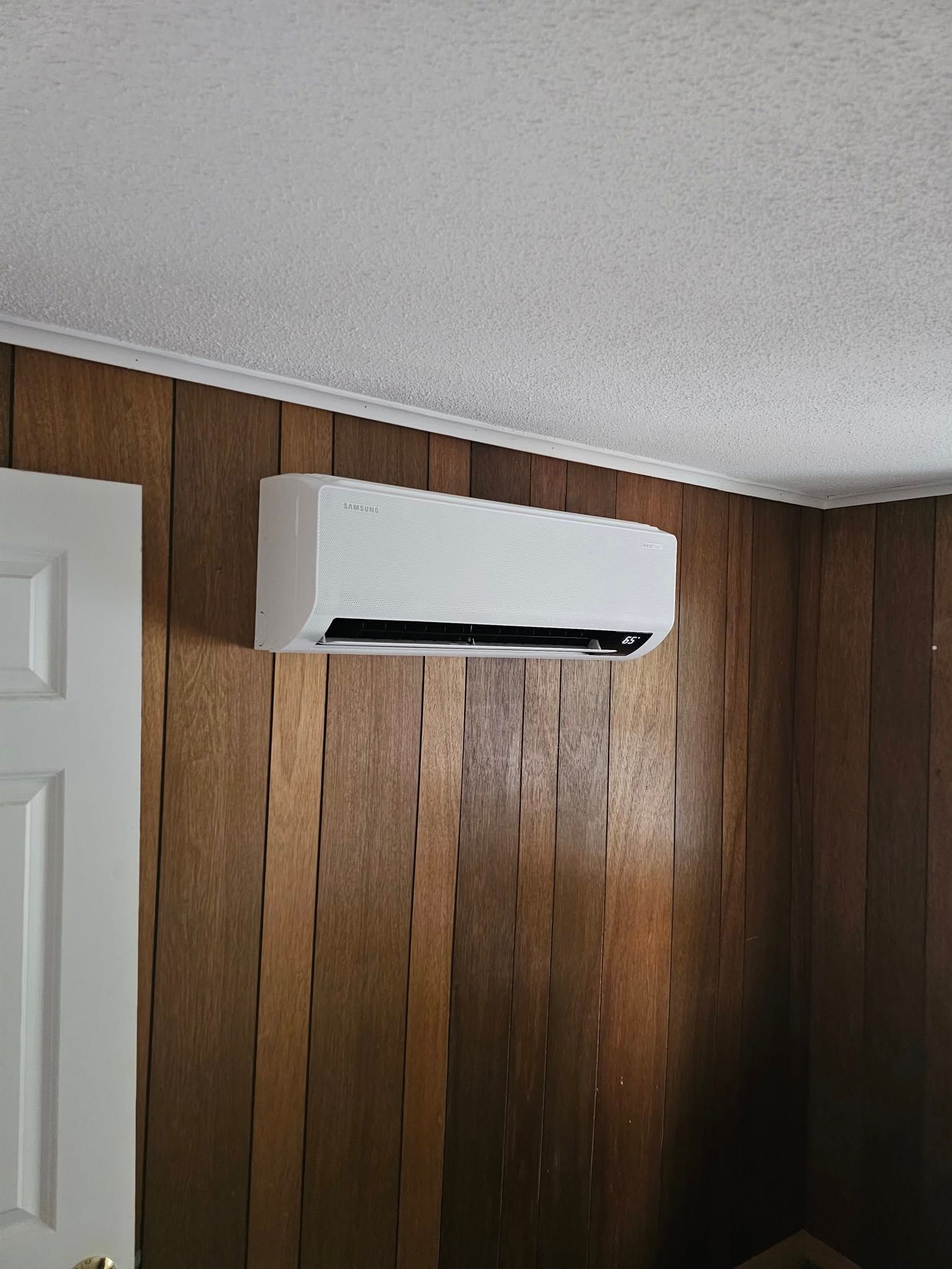 White air conditioning unit mounted on a wood-paneled wall, near a white door and ceiling.