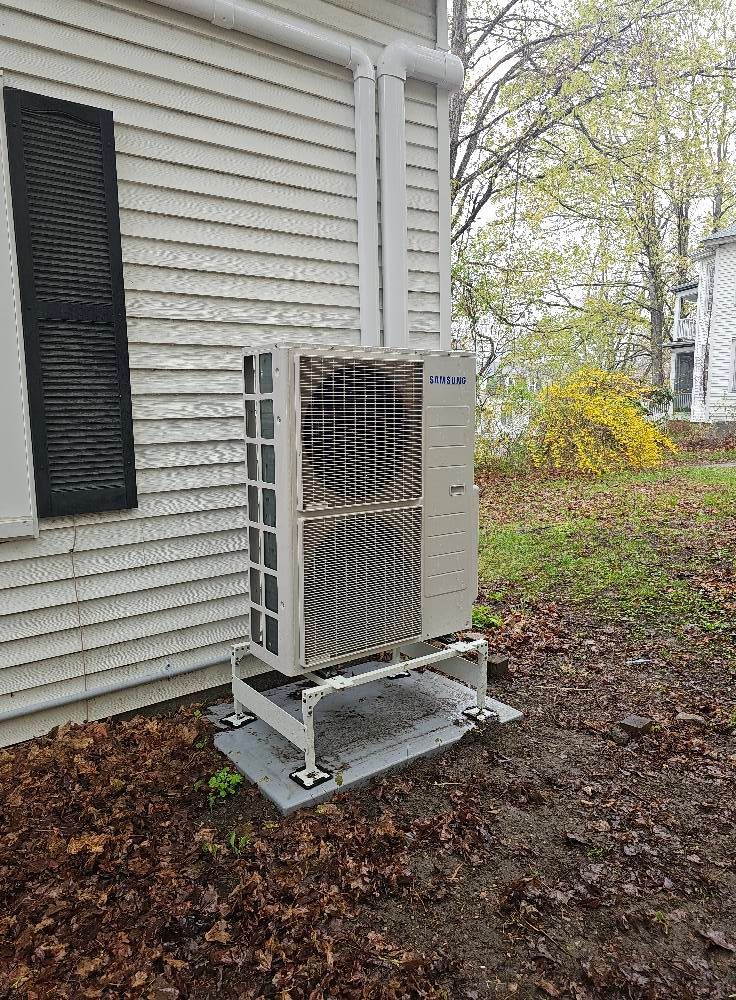An air conditioning unit on a raised platform next to a white house with a black shutter.