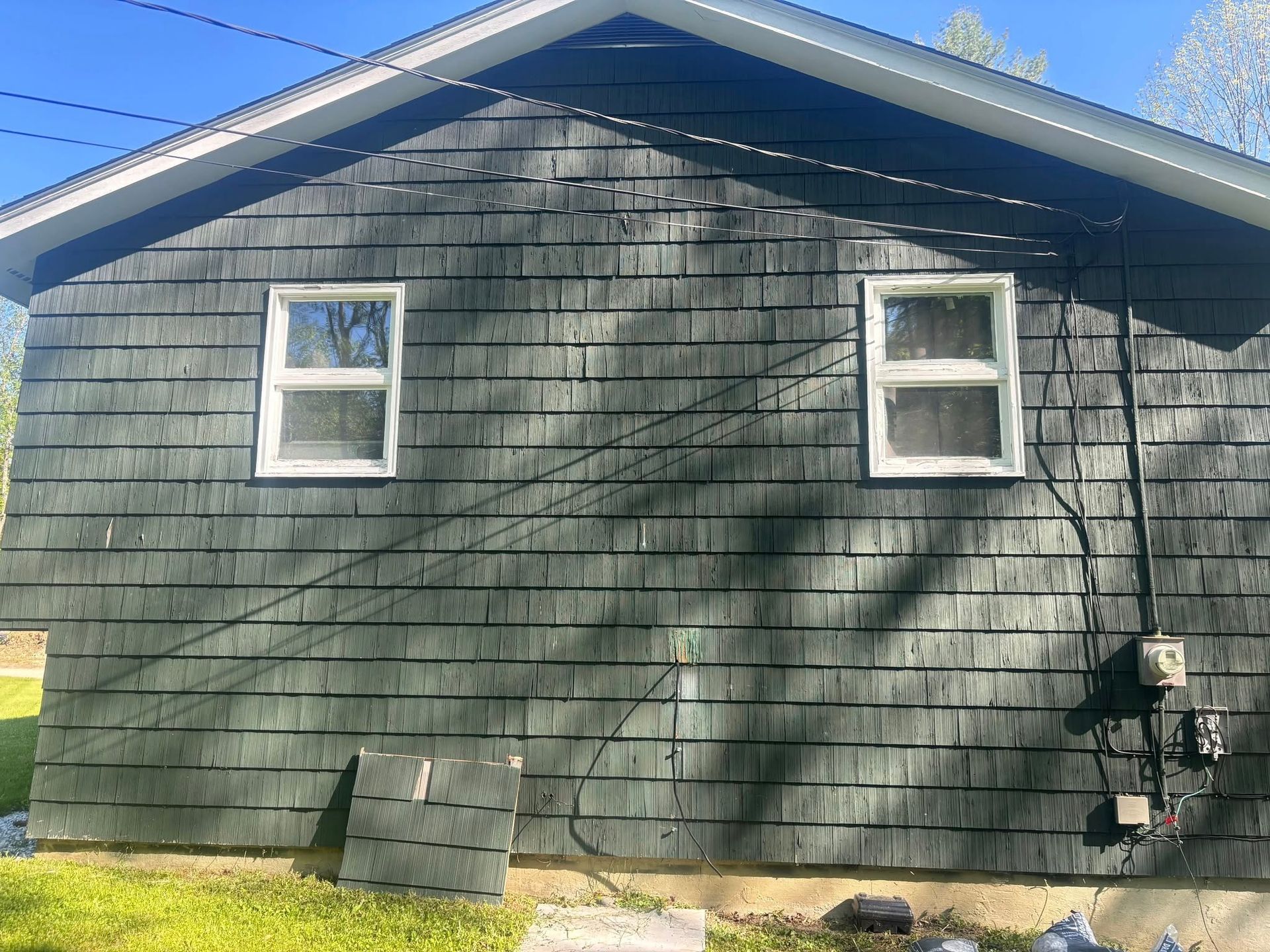 Dark-shingled house with two windows and white frames; electrical box on the right.