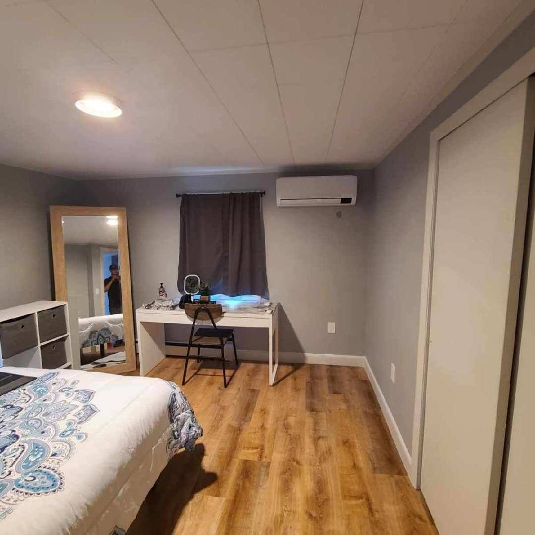 Bedroom with wooden floor, white desk, large mirror, bed, closet, and air conditioner. Gray walls and dark curtains.