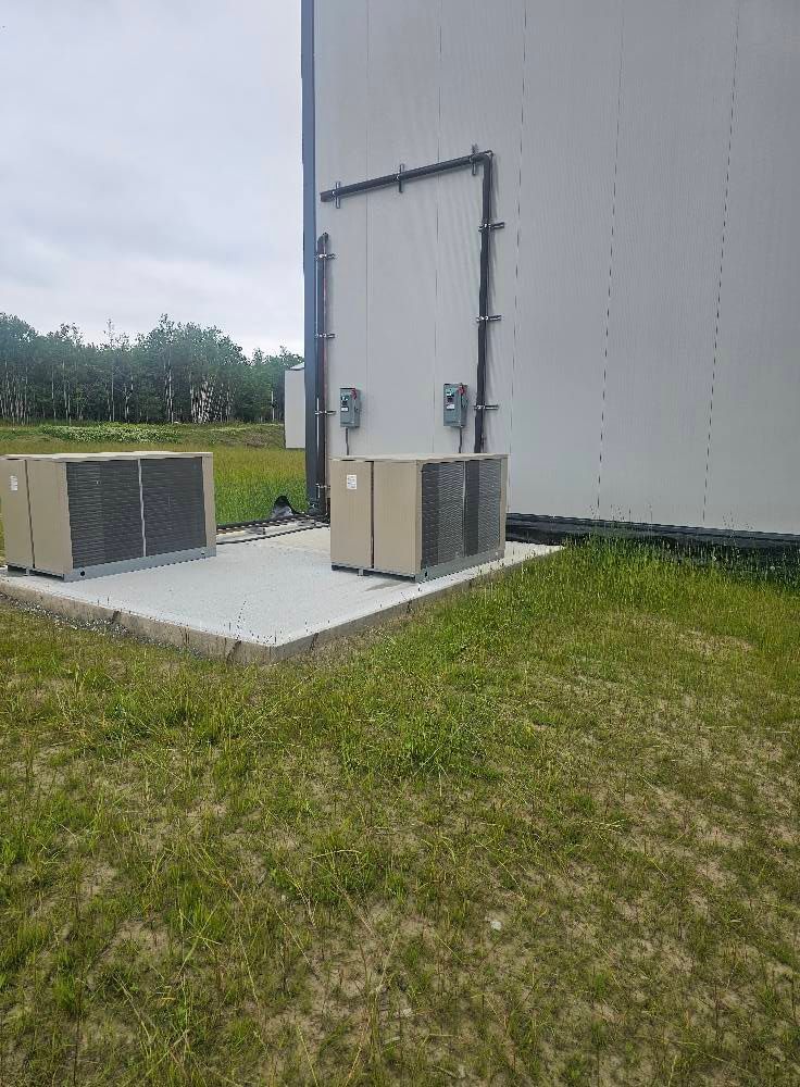 Two air conditioning units sit on a concrete pad outside a building with a door and conduit.