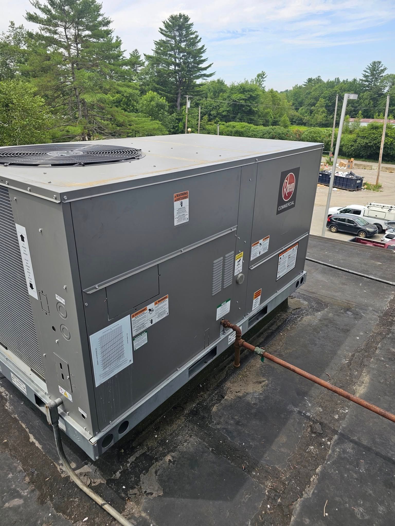 Large gray HVAC unit on a black roof, outdoors with trees and vehicles in the background.
