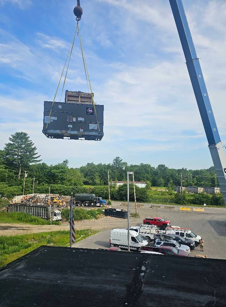 Crane lifting large, rectangular industrial equipment outdoors on a sunny day. Cars are parked below.