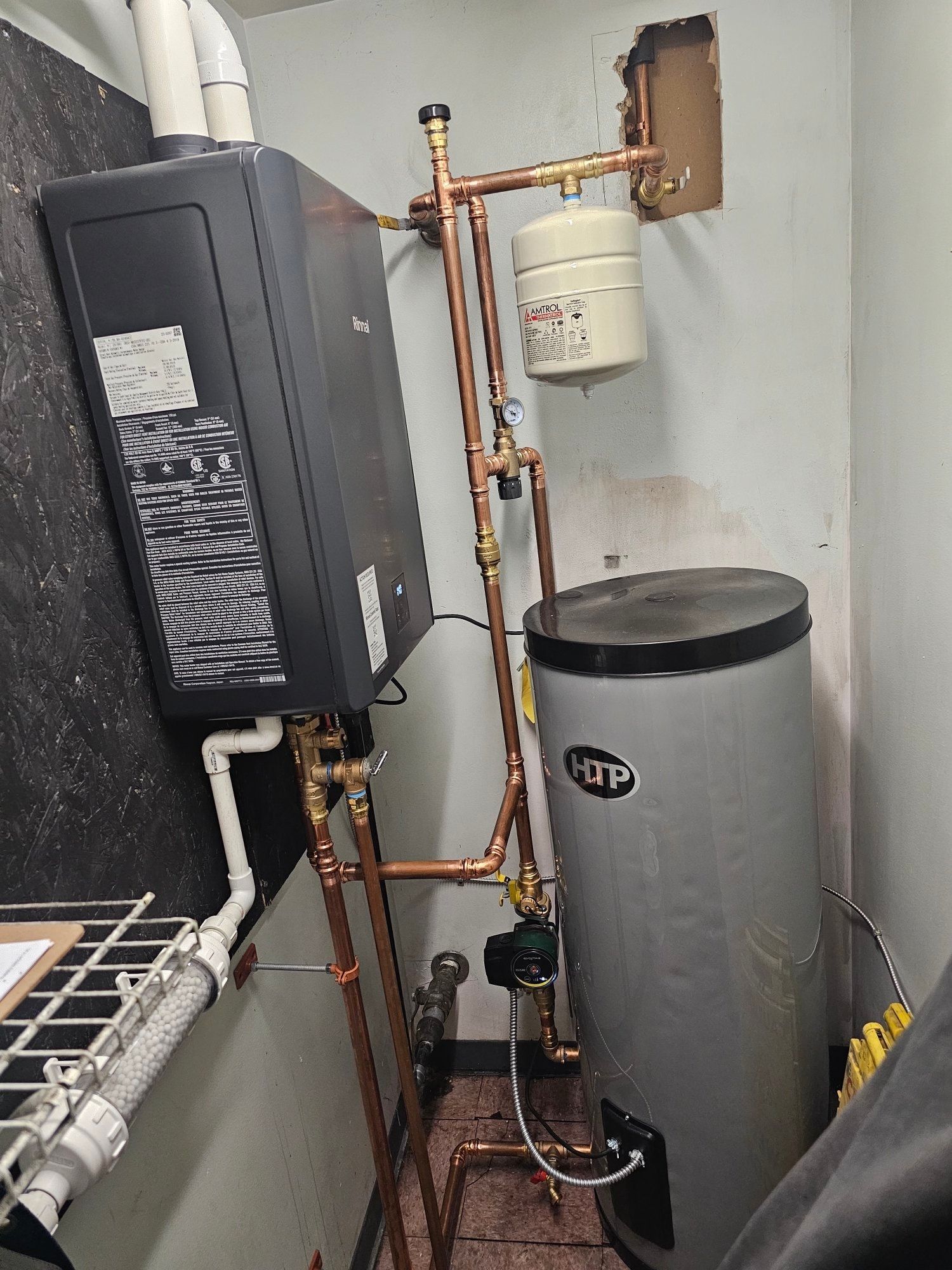 A wall-mounted black water heater with copper pipes, connected to a tall silver water tank in a small utility room.