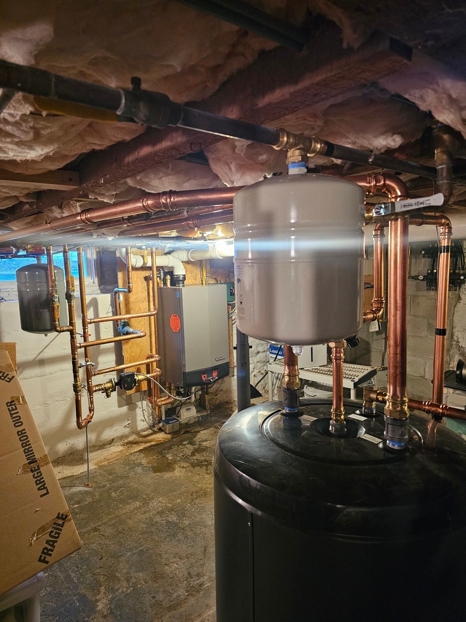 Copper pipes and tanks in a basement. The plumbing system has exposed copper pipes and large white and black tanks.