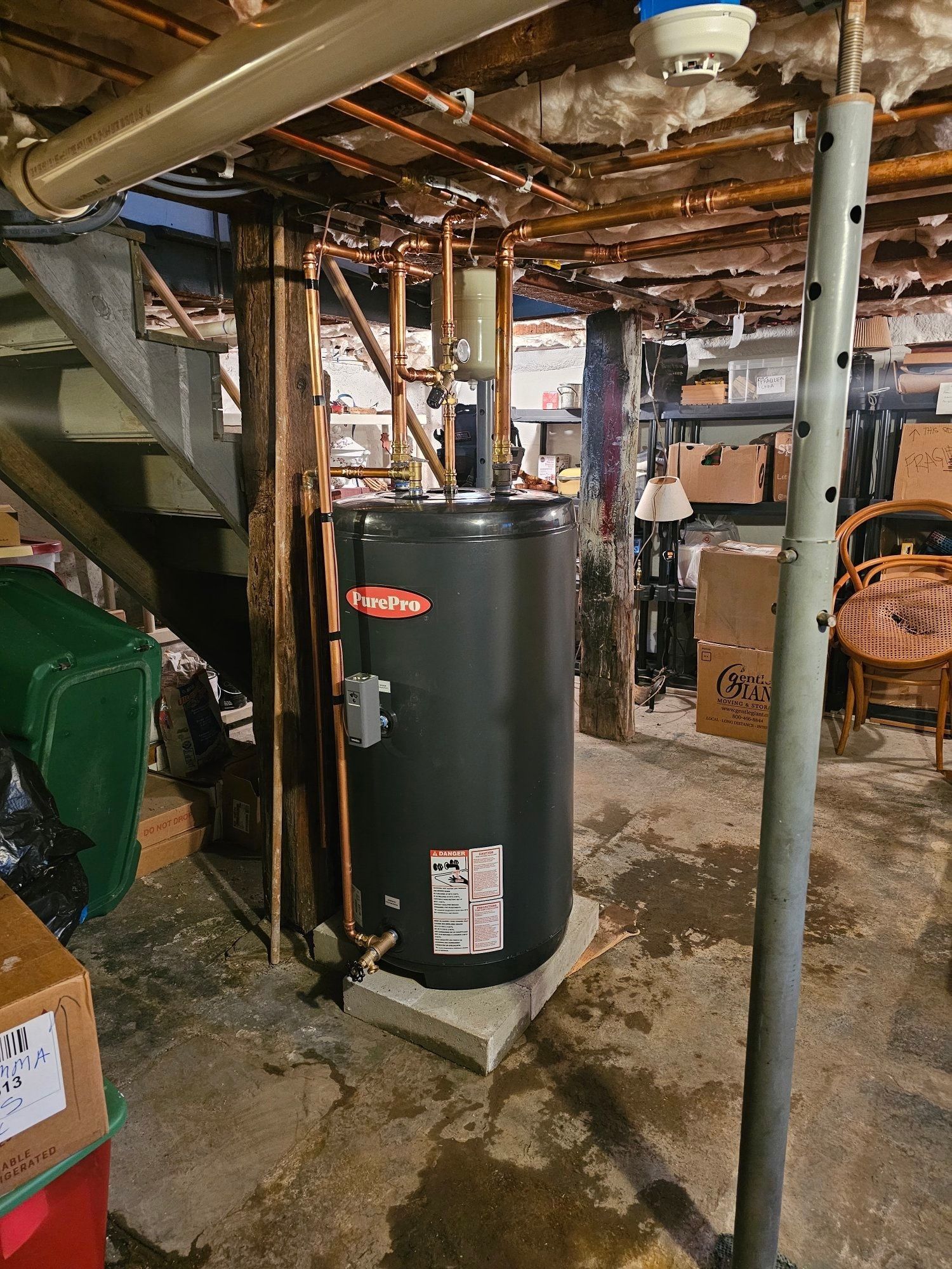 Black water heater in a basement with exposed pipes and stairs.
