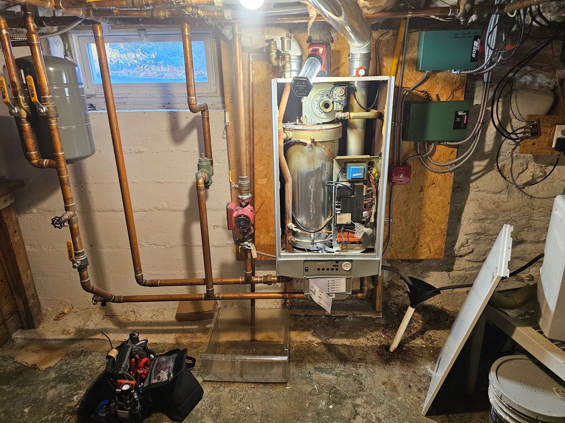 Furnace with open panels and exposed components, copper pipes, and water stains on a basement floor.