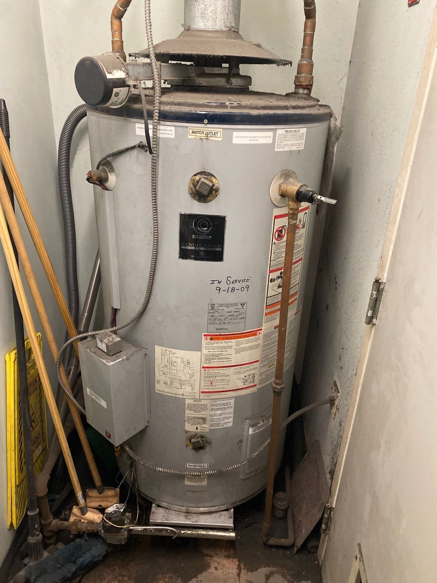 A gray water heater in a narrow, cluttered utility room with pipes and a broom.