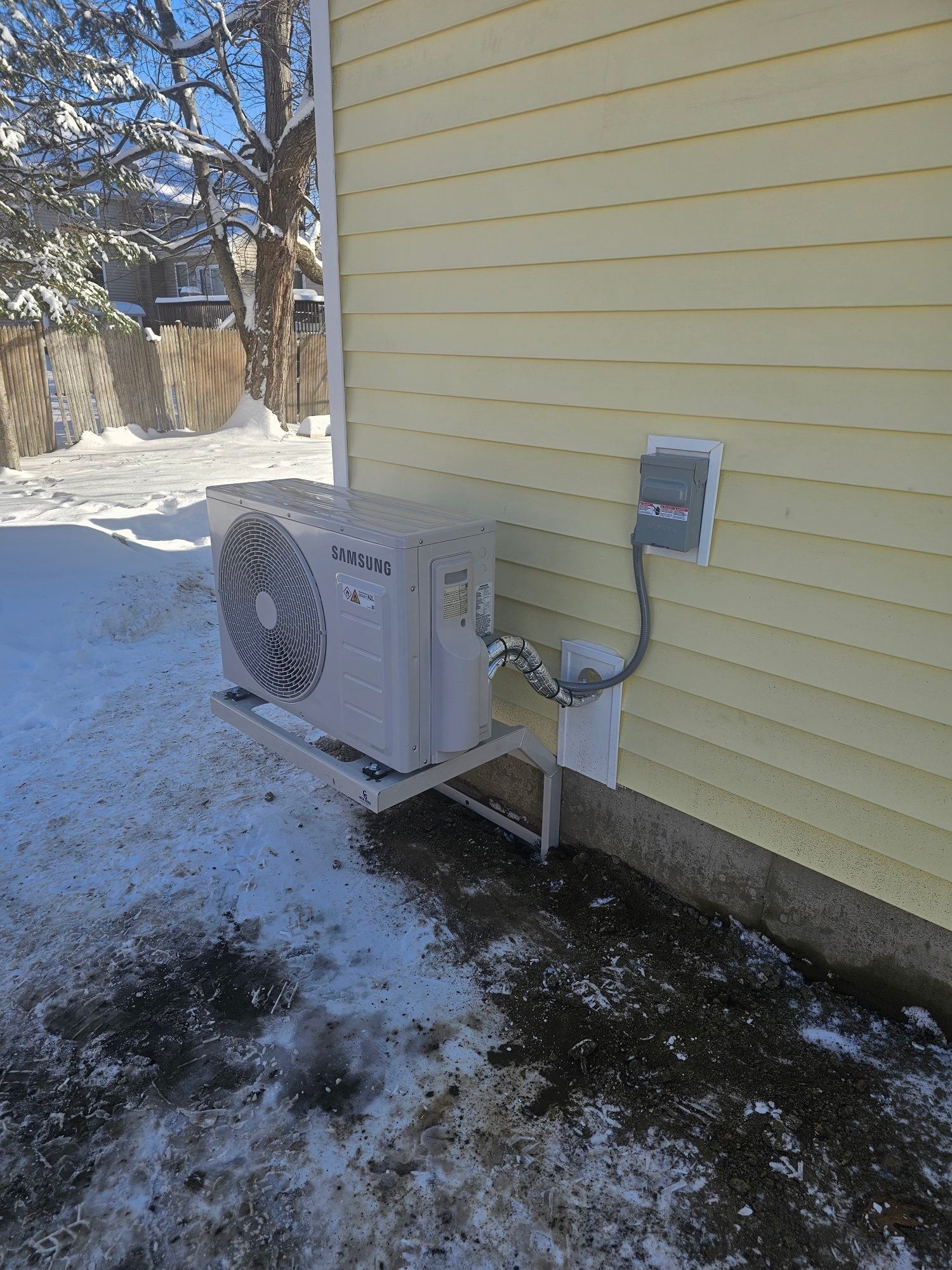 An outdoor white air conditioning unit mounted on a light-colored building in a snowy environment.