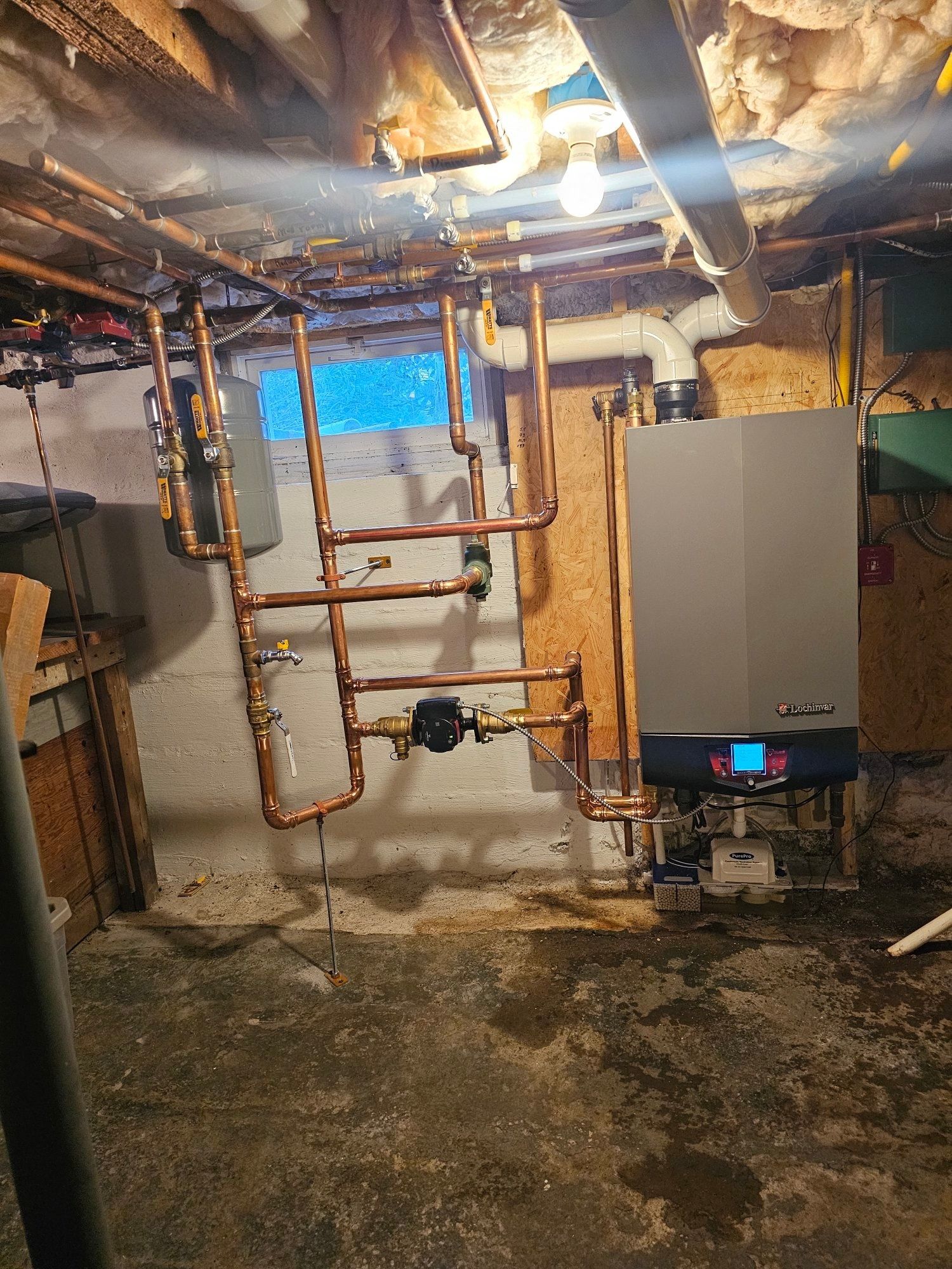 Copper pipes and a boiler in a basement, on a concrete floor.