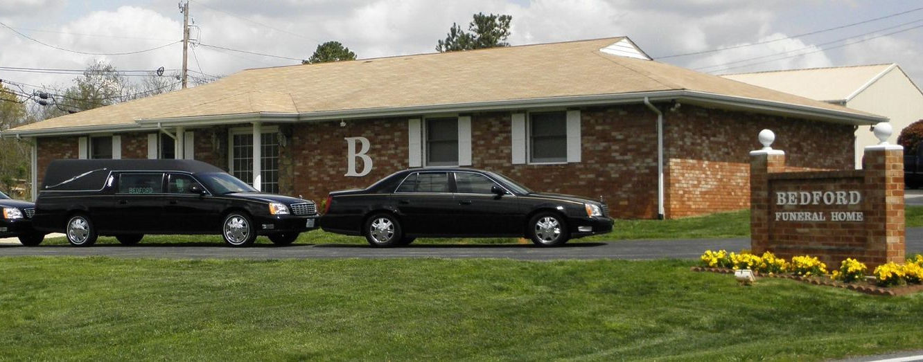 A hearse and a black sedan parked in front of the brick Redford Funeral Home building under a cloudy sky.