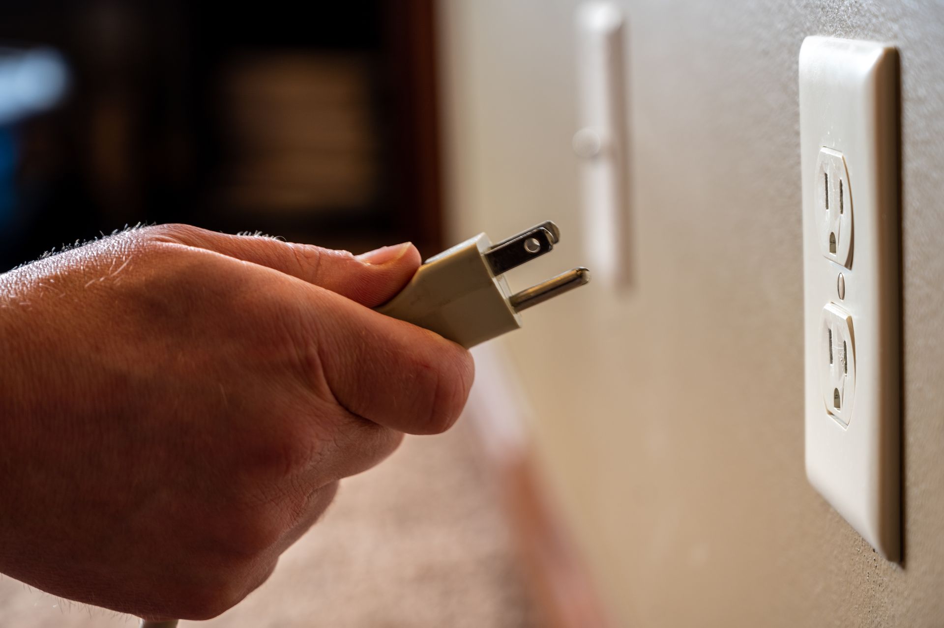 Hand plugging a cord into a white electrical outlet on a beige wall.