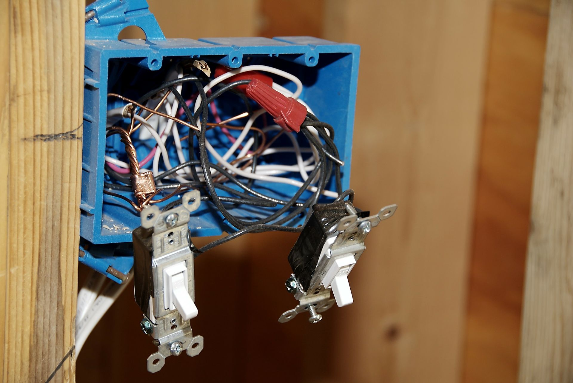 Blue electrical box with tangled wires and two attached light switches, mounted on a wooden wall.