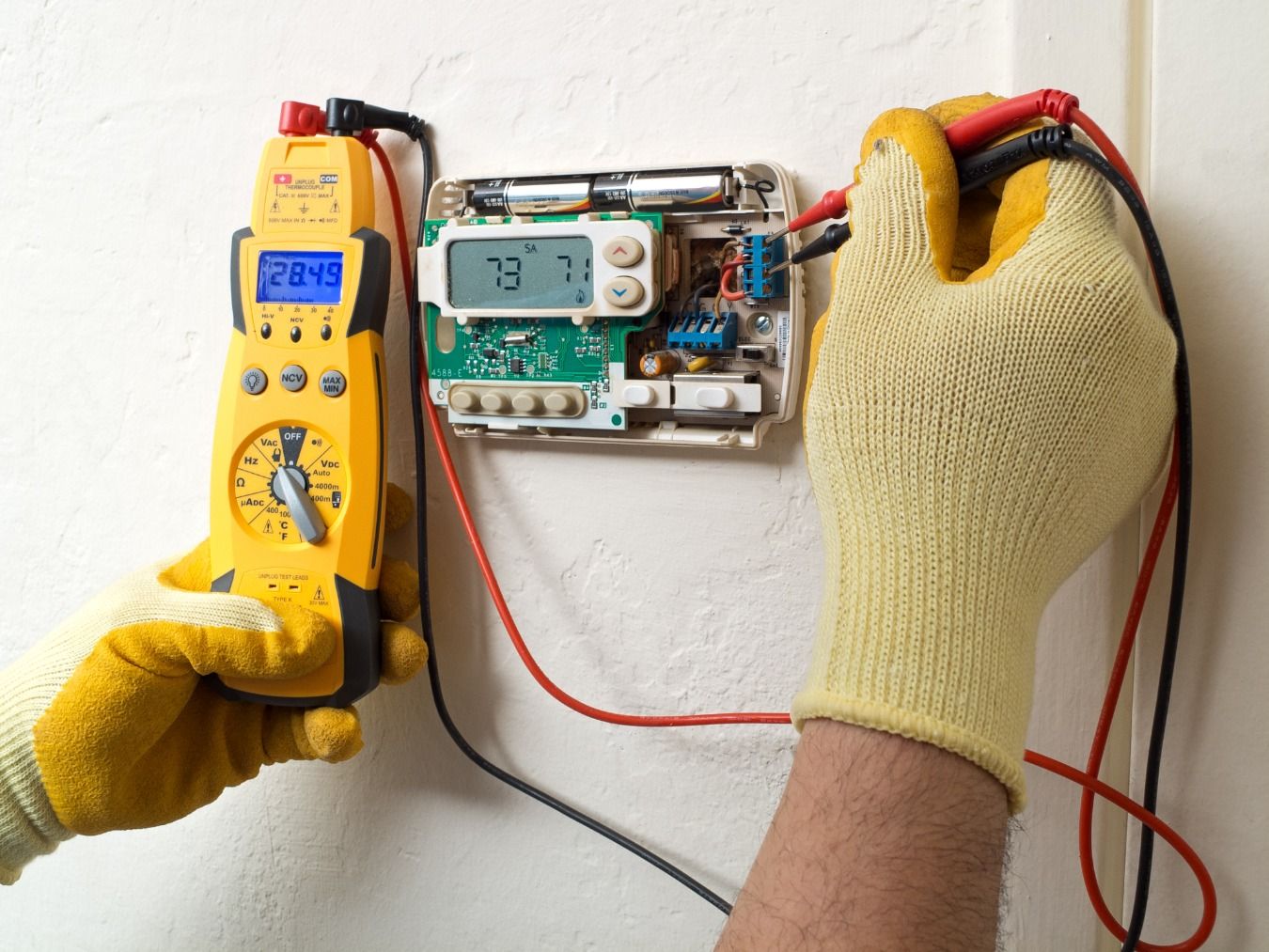 Hands wearing gloves testing a thermostat with a multimeter. Yellow meter displays 28.49.