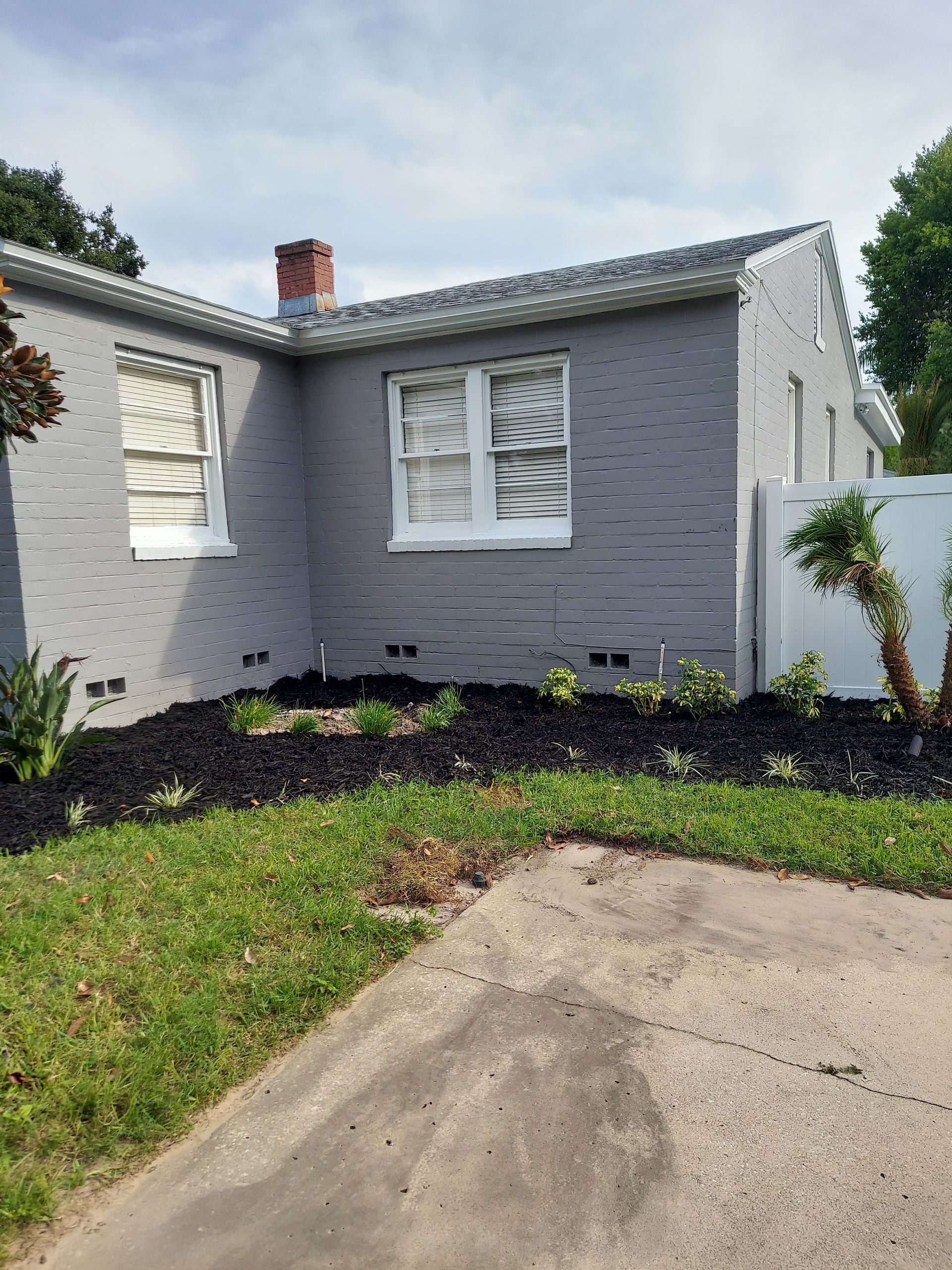 A gray brick house with a white fence and a driveway in front of it.
