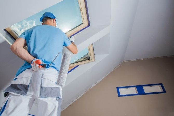 Worker painting a ceiling window frame in a room with blue painter’s tape on the wall