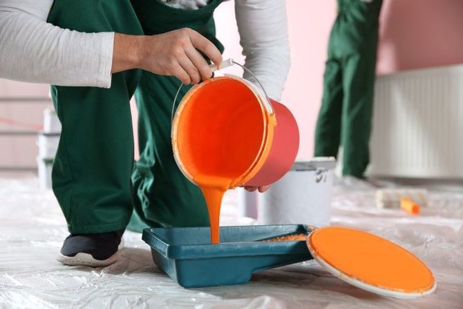 Person pours bright orange paint into a tray on a plastic-covered floor, preparing to paint indoors.