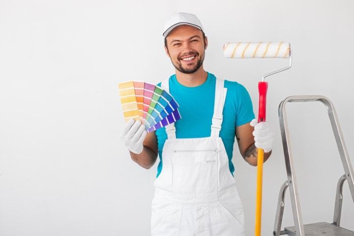 Painter holding color swatches and paint roller beside ladder, smiling in white overalls and blue shirt