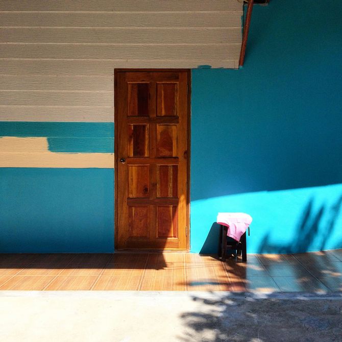 Wooden door in a blue wall beside a small stool with a pink cloth in sunlight.