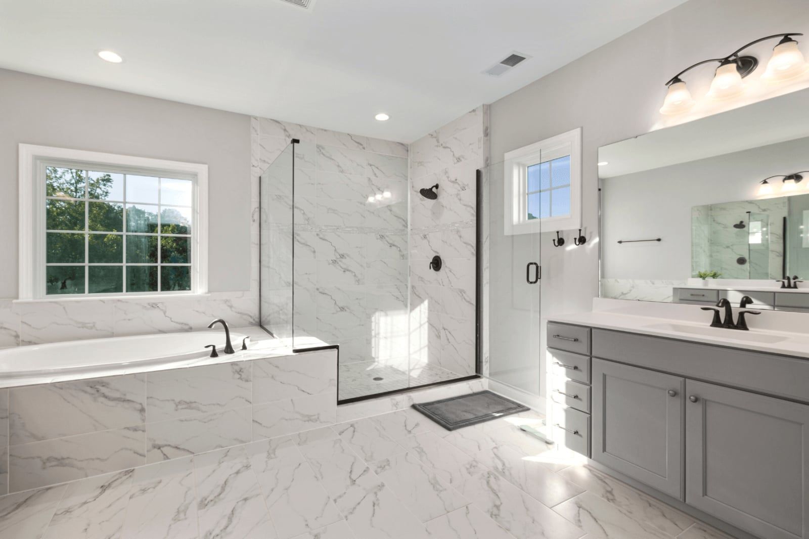 Bright white luxury bathroom with marble tub, glass shower, gray vanity, and large window.In Cincinnati, OH.