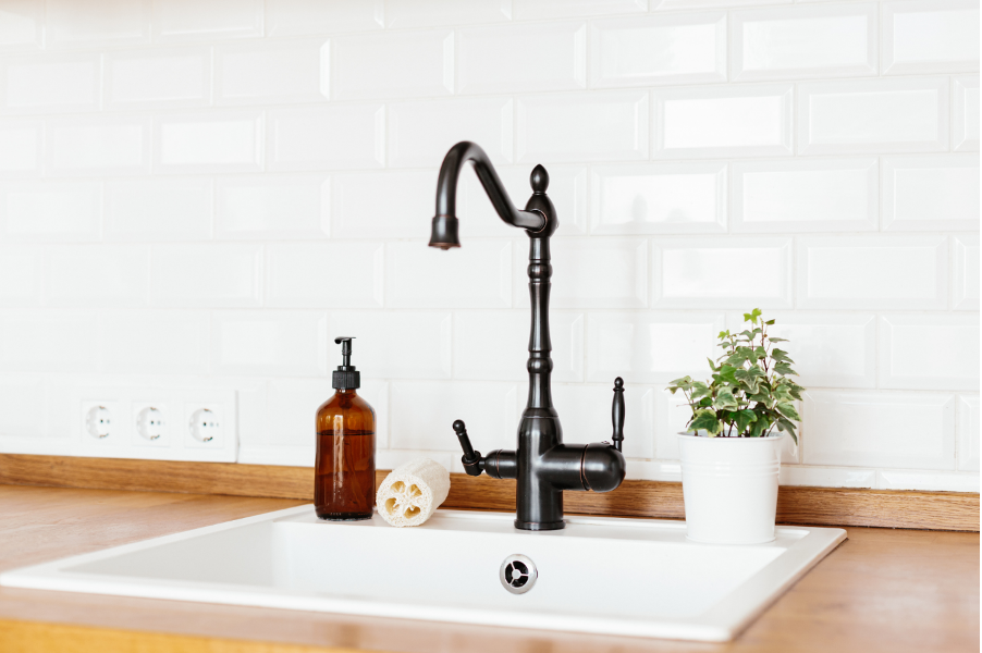 A kitchen sink with a black faucet , soap dispenser , sponge and potted plant.