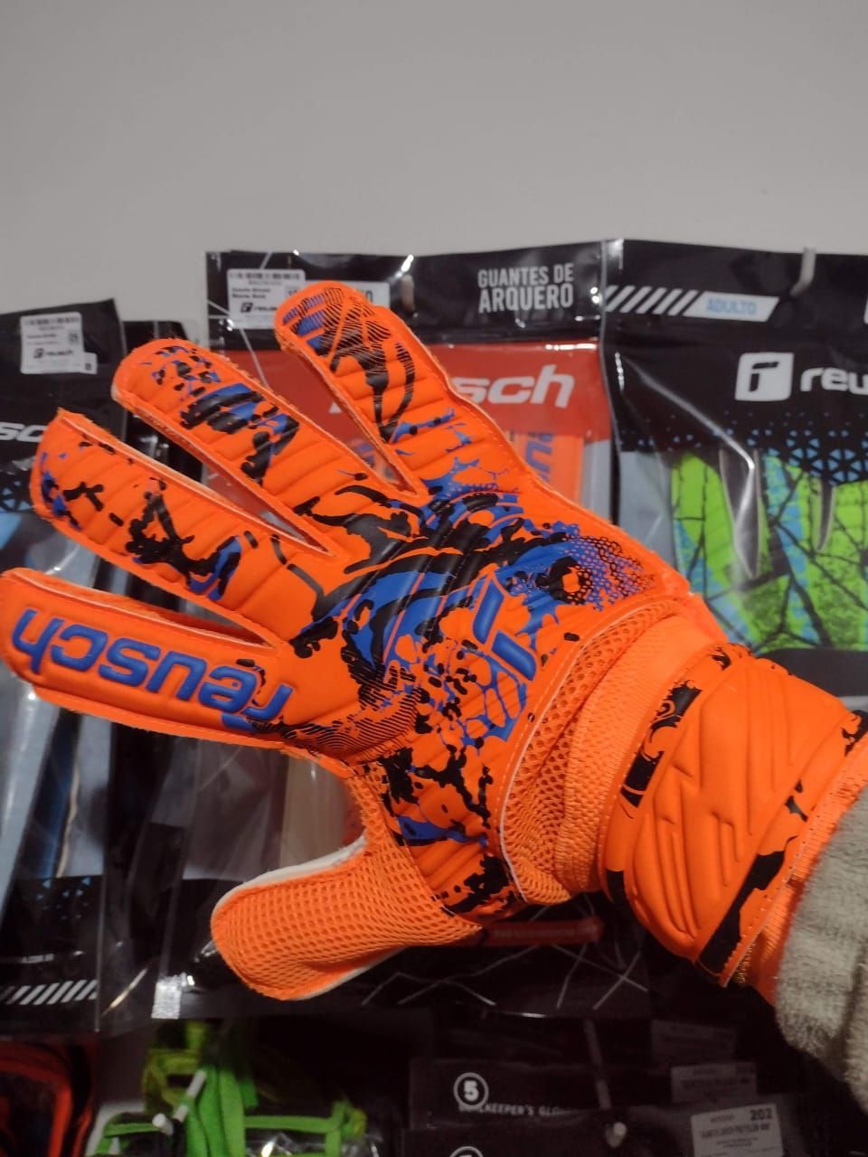 Par de guantes de portero Reusch naranjas y blancos, uno con la palma y el otro con el dorso pintados con un diseño de salpicaduras de pintura azul.