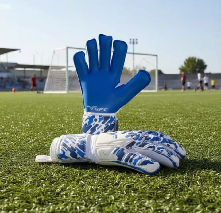 Guantes de portero azules y botas de fútbol blancas y azules en un campo de fútbol con una portería al fondo.