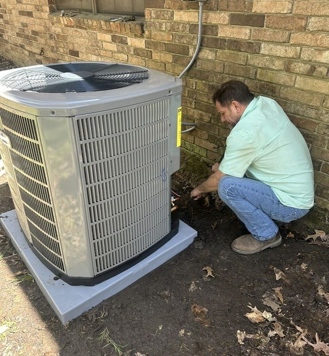 Technician crouches beside an outdoor air conditioner unit, inspecting wiring by a brick wall.