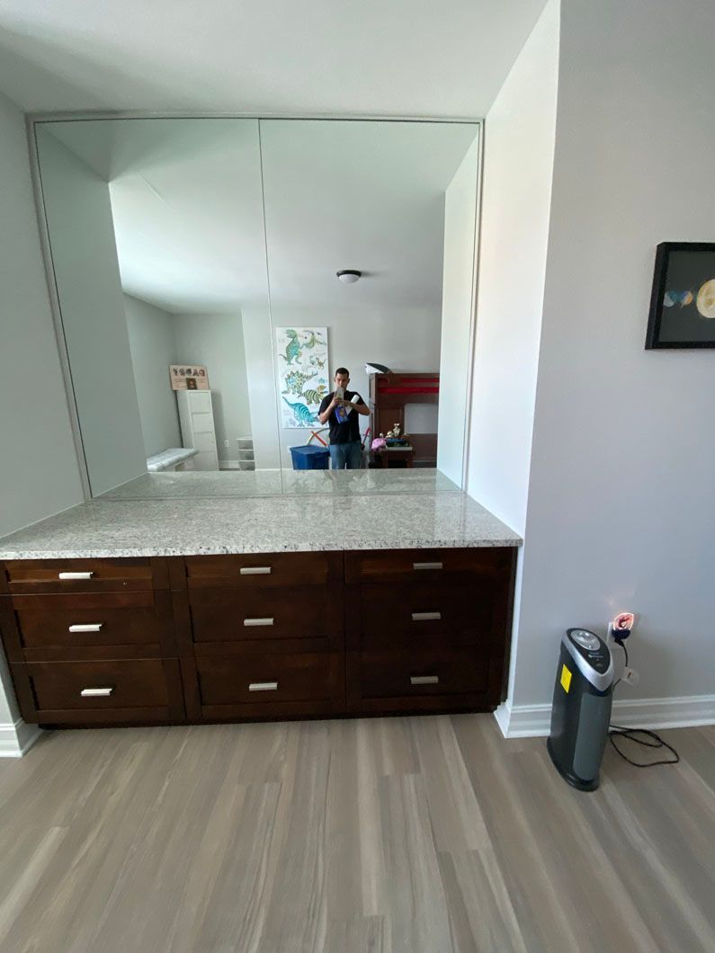 Built-in wooden dresser with granite countertop, large mirror, and person reflected in it. Gray walls and flooring.