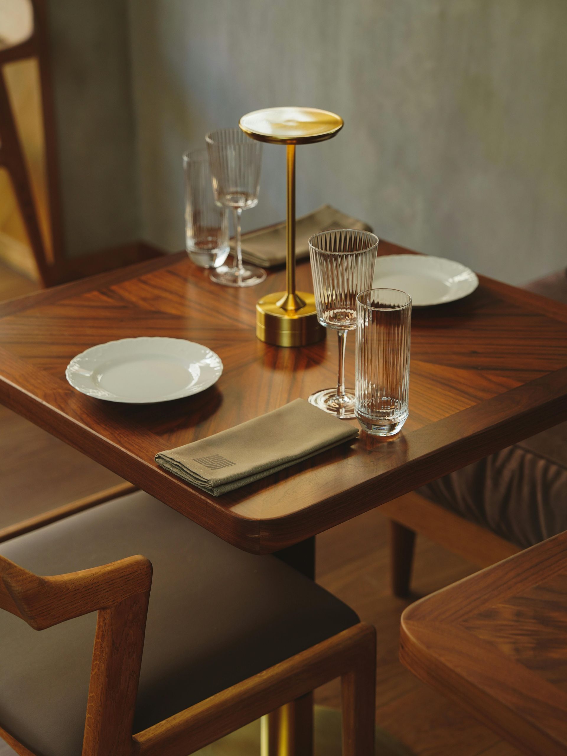 Square wooden table set for dining with glasses, plates, and a lamp.