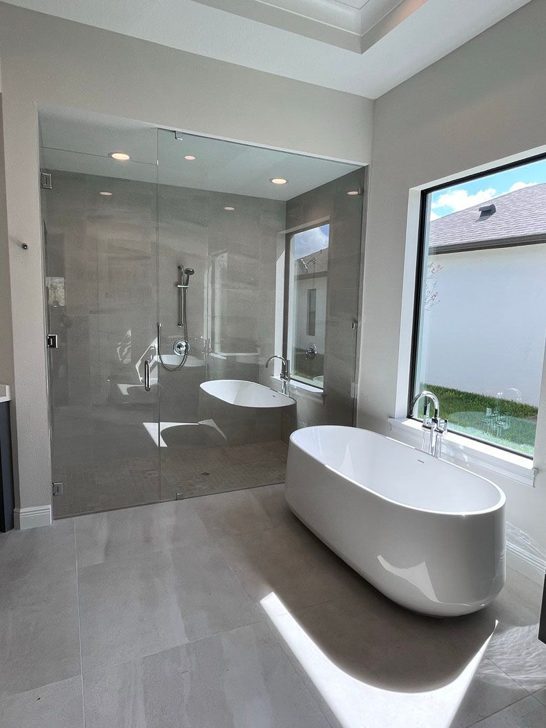 Modern bathroom with gray tiled walls and flooring. Large glass shower enclosure, freestanding white bathtub.