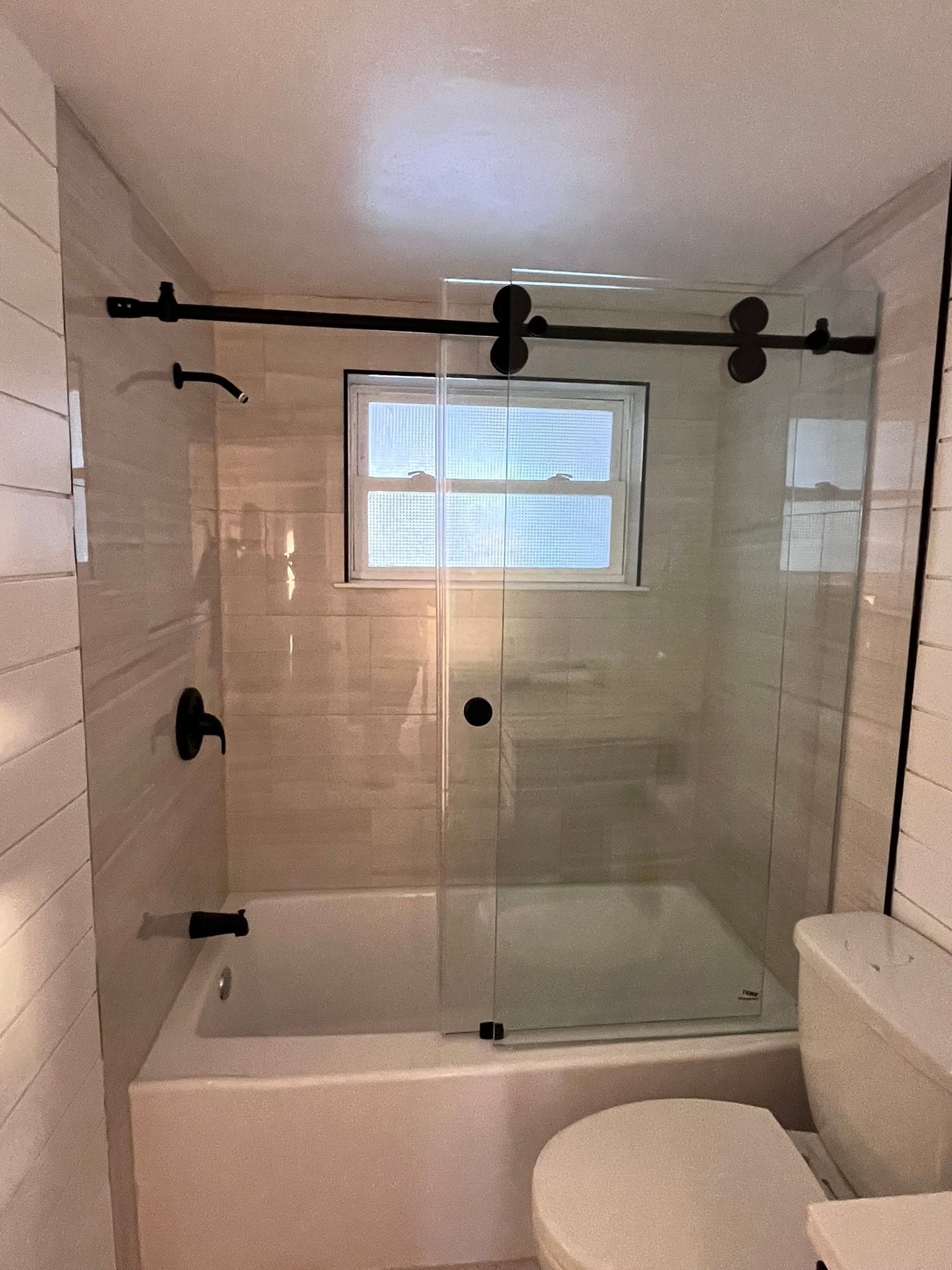 Bathroom with a white bathtub, glass shower door, and window. Black hardware and white tiles.