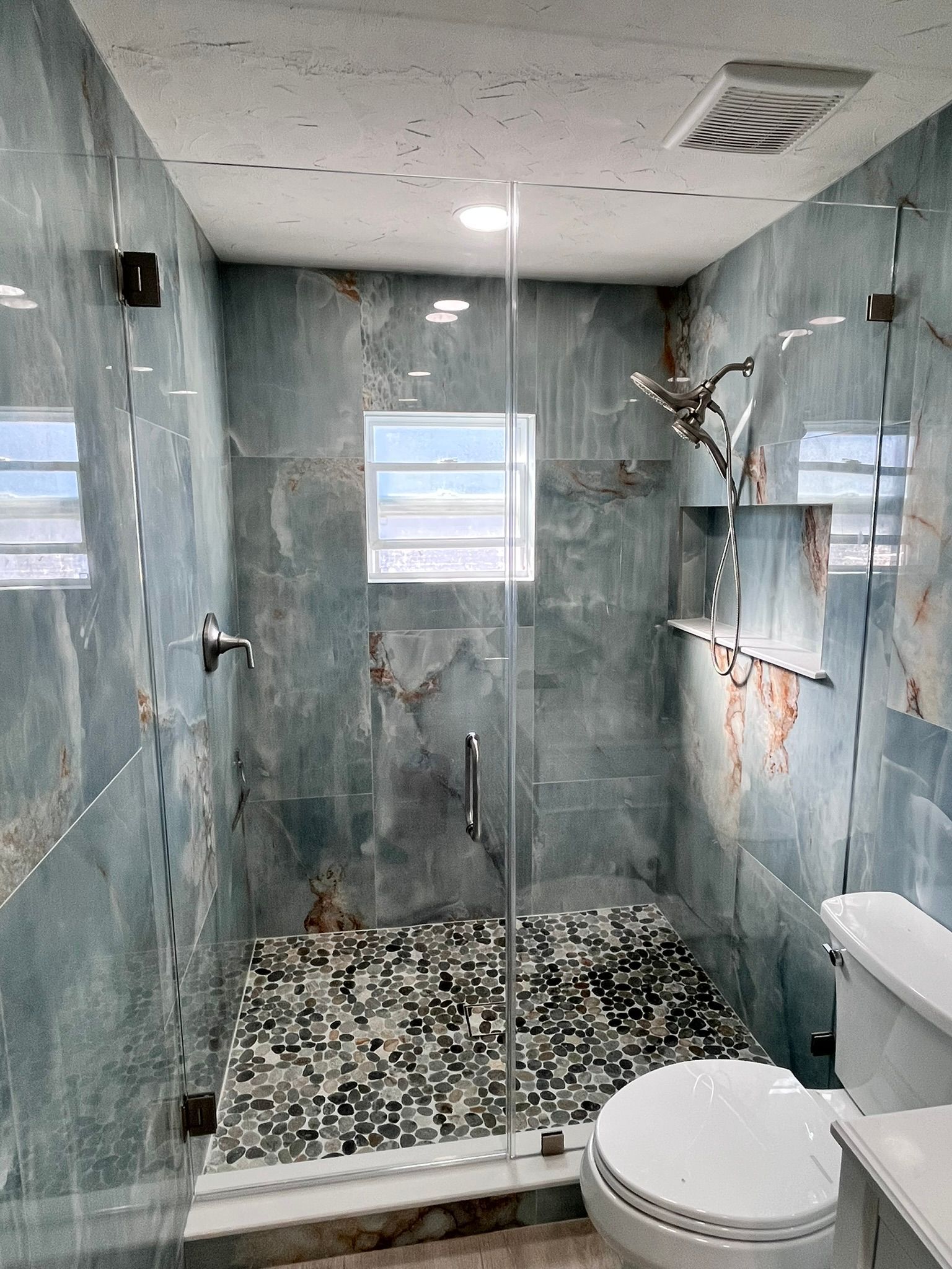 Blue-tiled shower with a glass door. Pebble floor, white toilet, and recessed shelves.