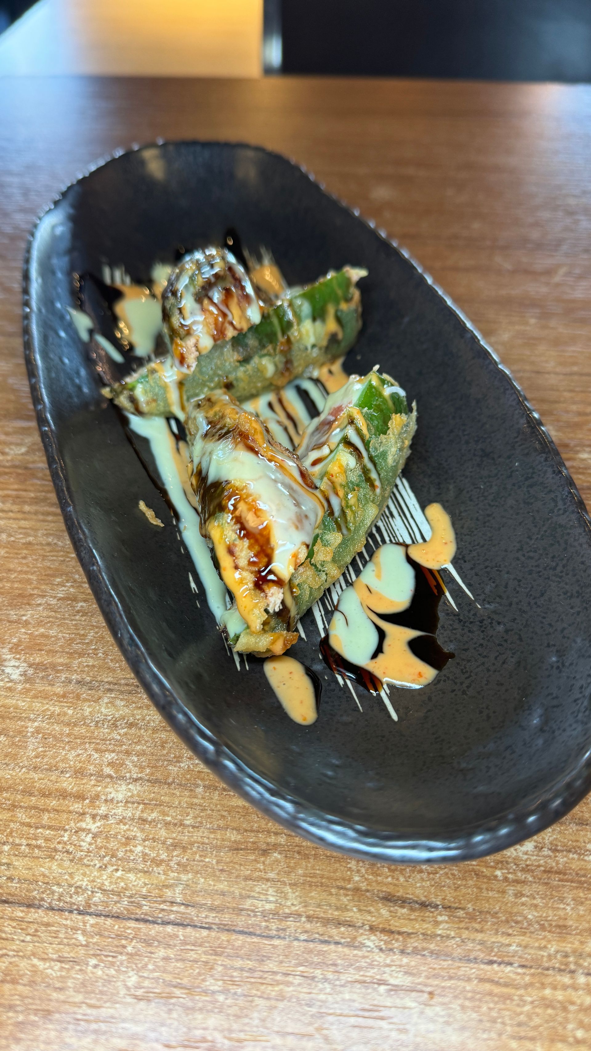 Sushi roll slices on black plate, drizzled with sauces, on a wooden table.