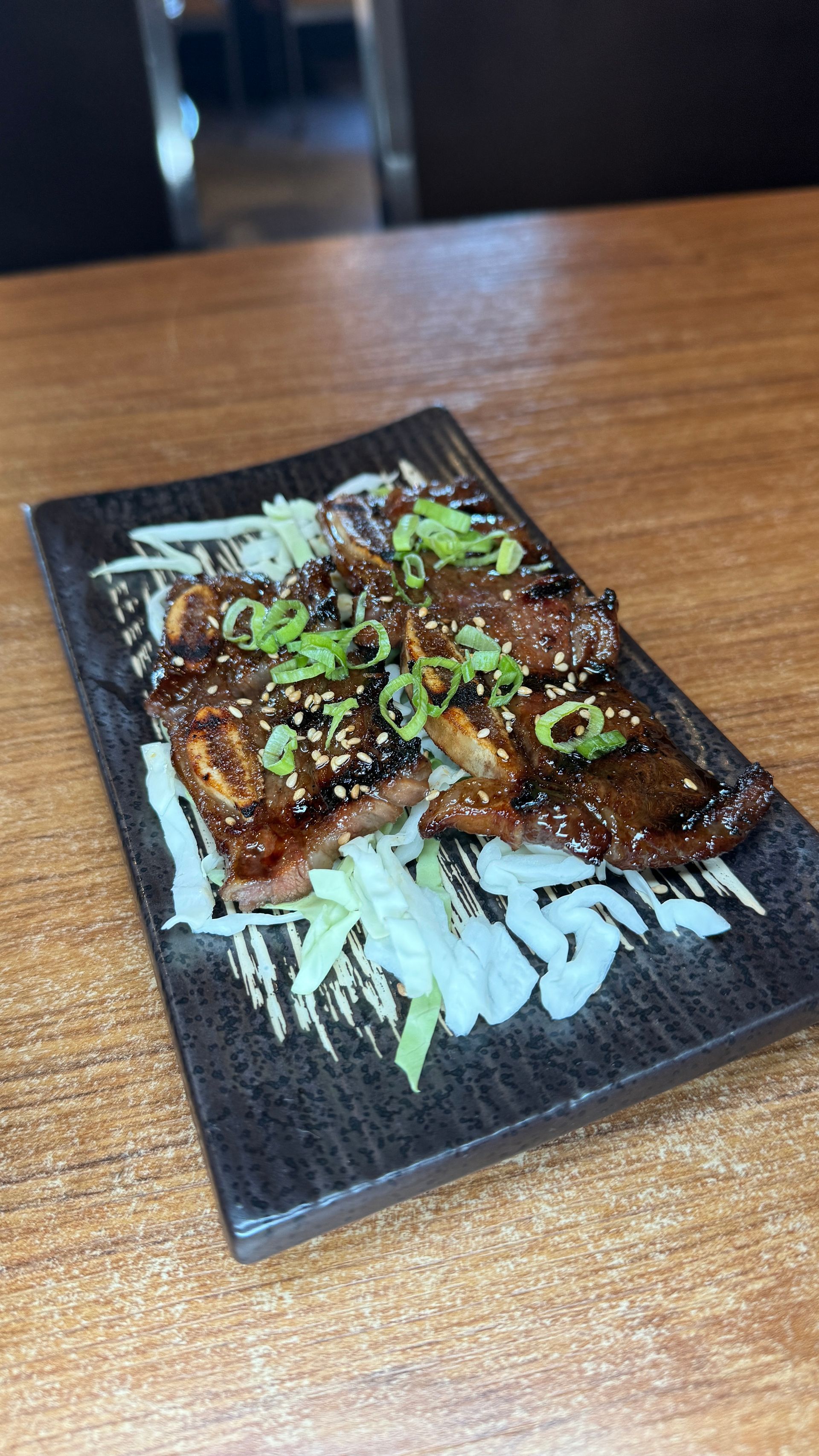 Grilled meat dish on a rectangular plate with shredded cabbage, sesame seeds, and green onions.
