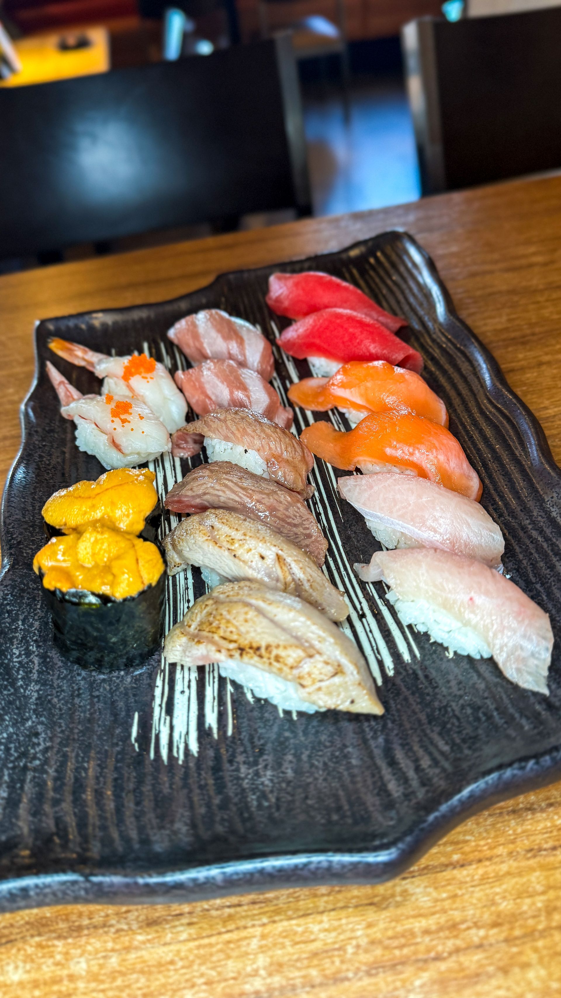 Sushi platter: various nigiri pieces, including salmon, tuna, shrimp, and sea urchin, on a black, textured plate.