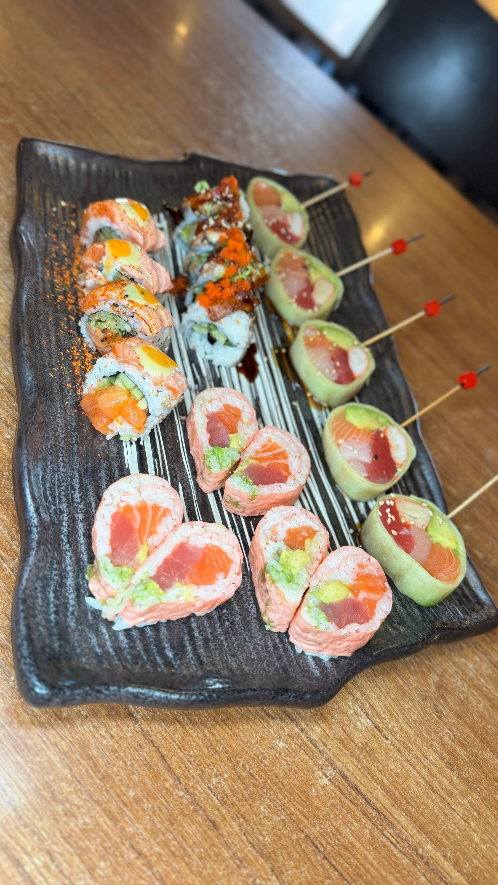 Sushi platter with various rolls, including heart-shaped and skewers, on a dark, textured plate.