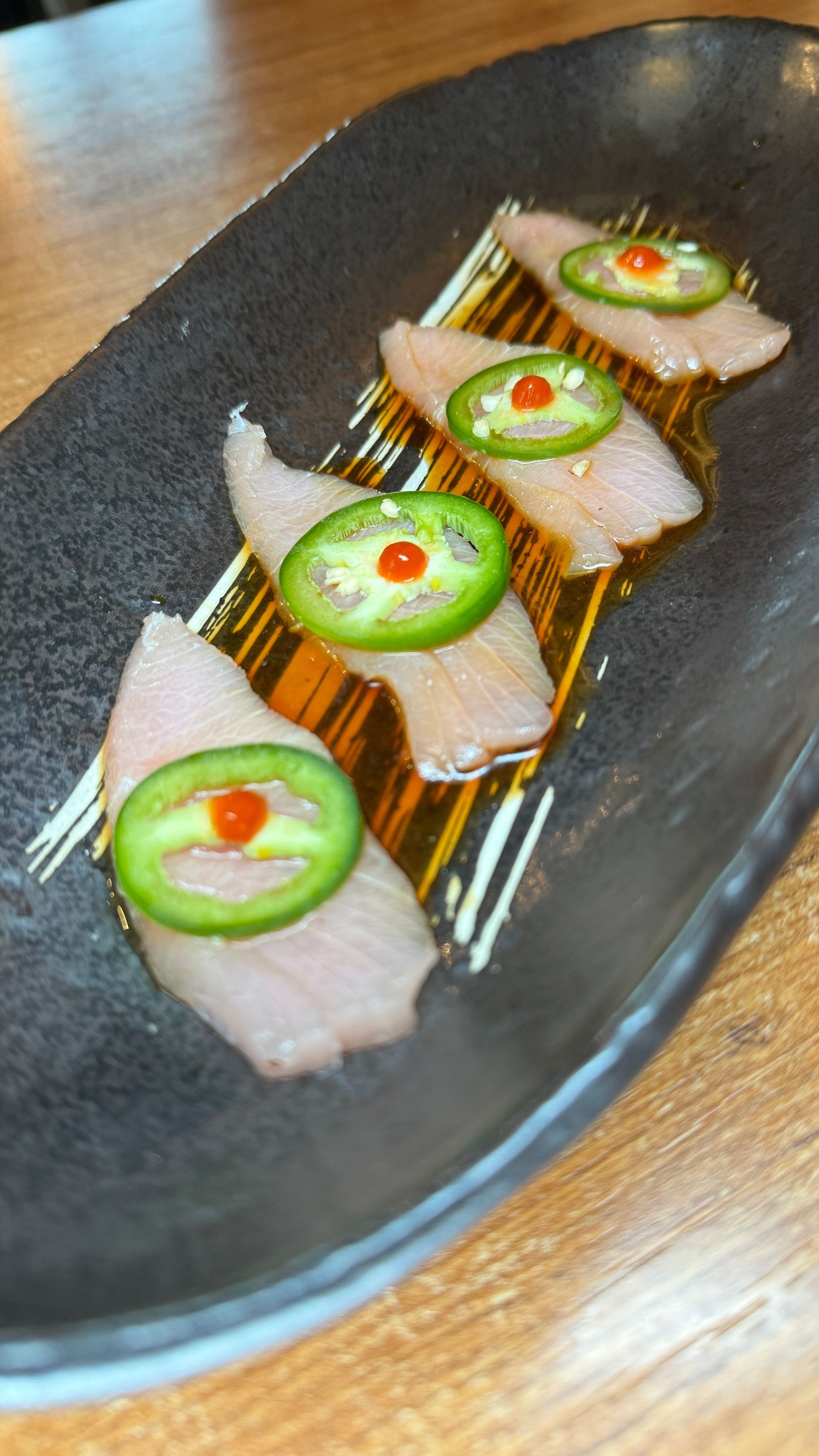 Sushi appetizer on a black plate: raw fish slices with jalapeños and red sauce, drizzled with sauce.