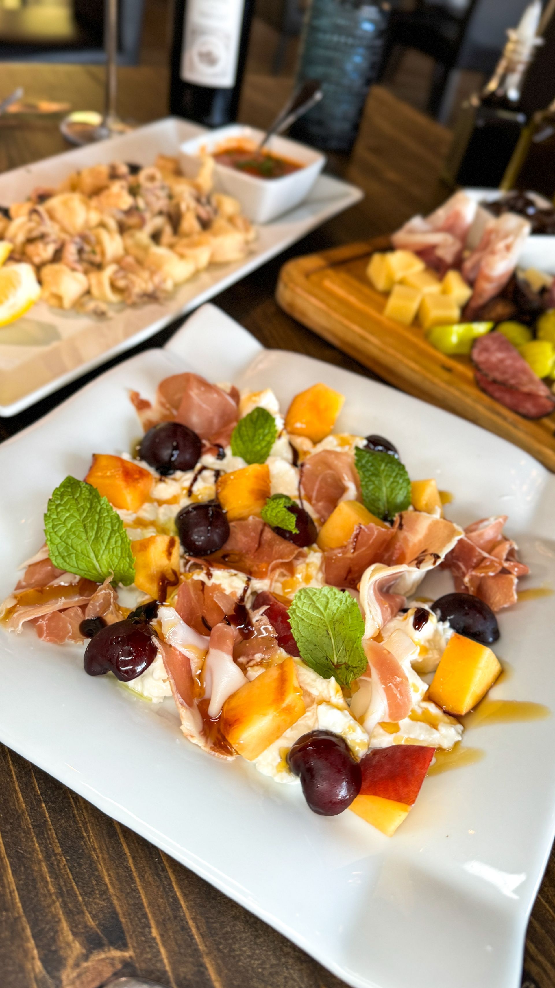 A plate with prosciutto, fruit, and cheese, other dishes, and wine bottles on a table.