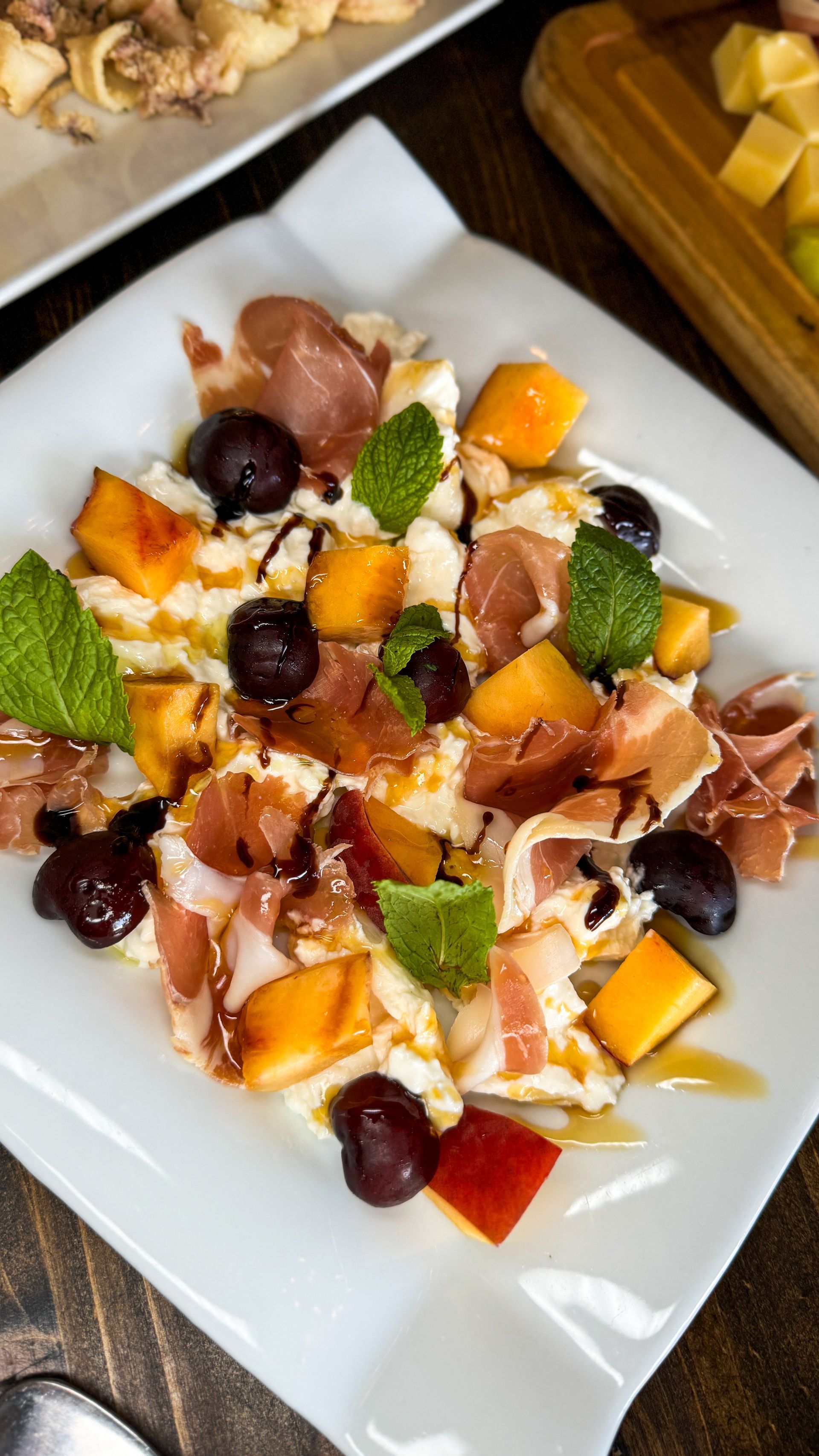 Salad with prosciutto, cantaloupe, grapes, cheese, mint, and balsamic glaze on a white plate.