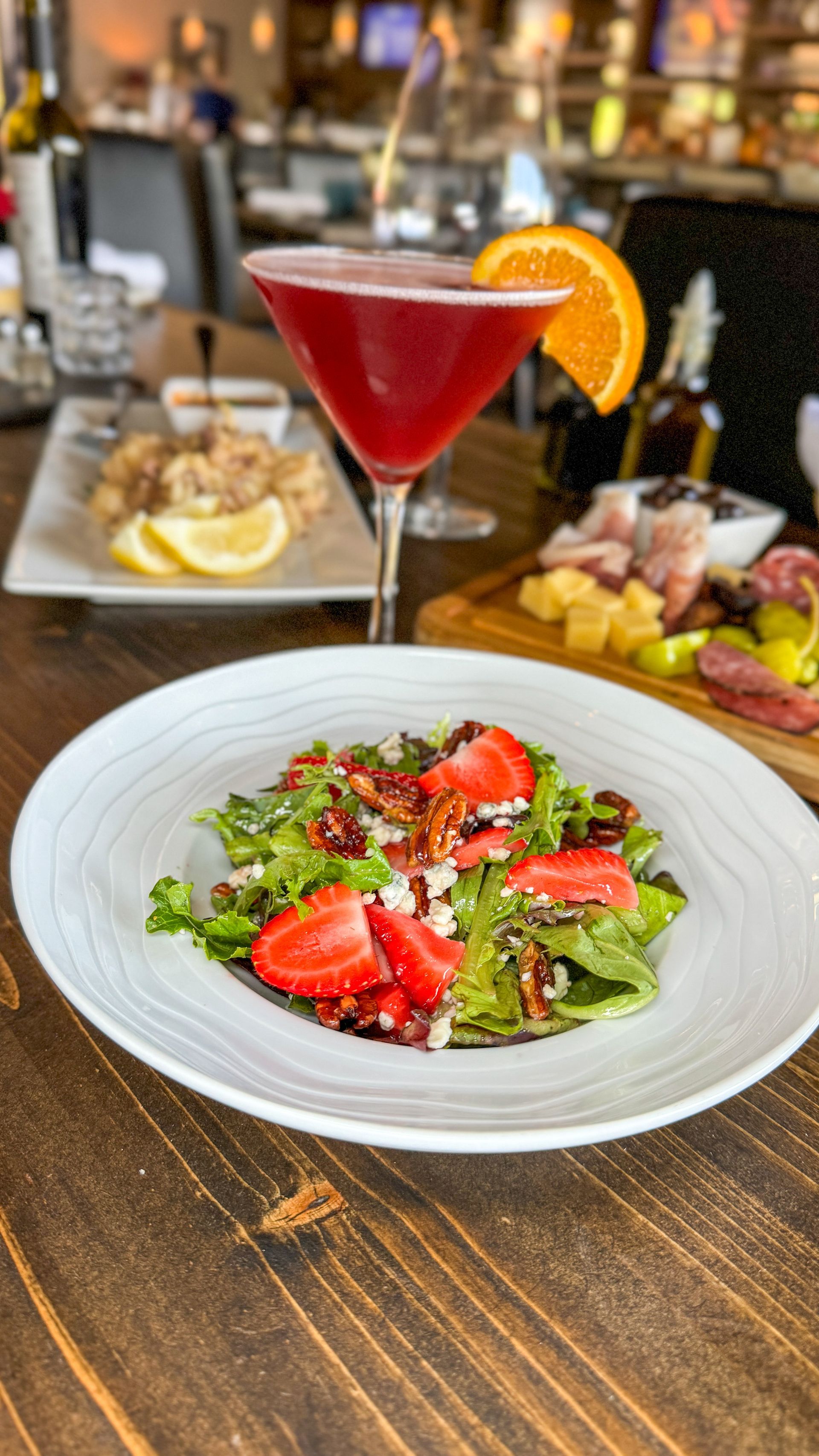 A strawberry salad, cocktail, and charcuterie board on a wooden table in a restaurant.