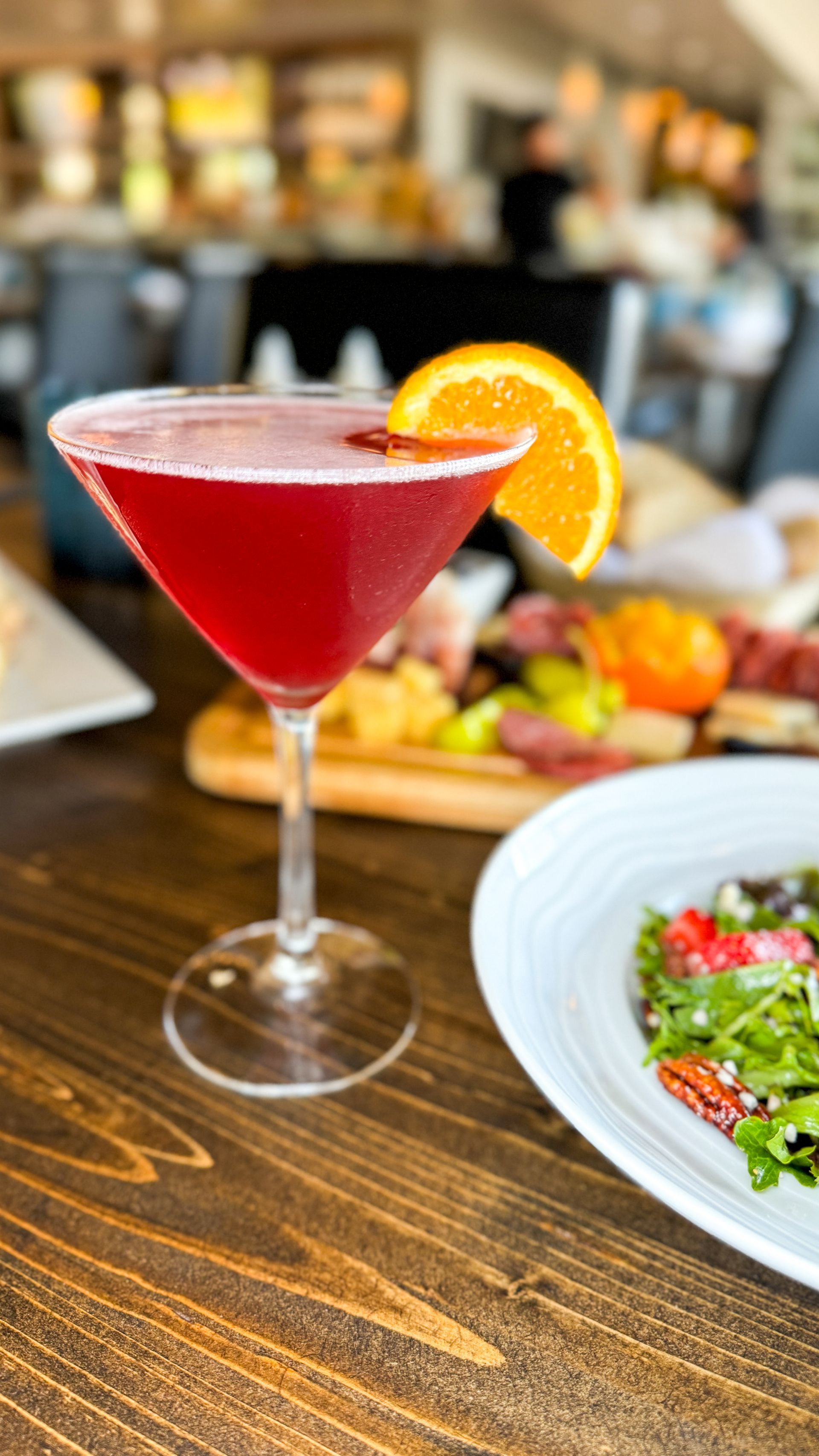 Cosmopolitan cocktail with orange slice garnish on wooden table, charcuterie board and salad in background.