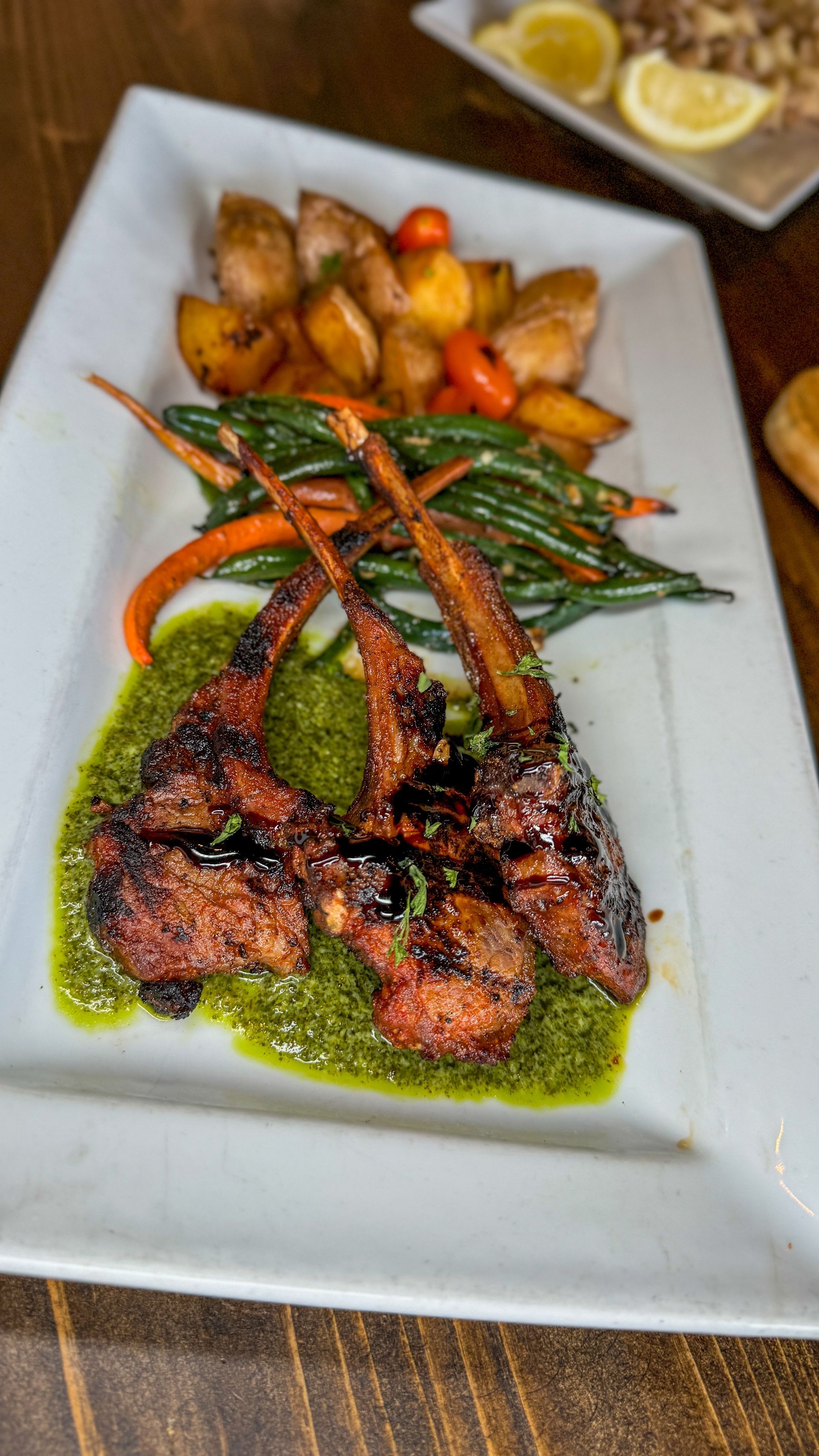 Grilled lamb chops with pesto, roasted potatoes, green beans, carrots. Plated on white rectangle.