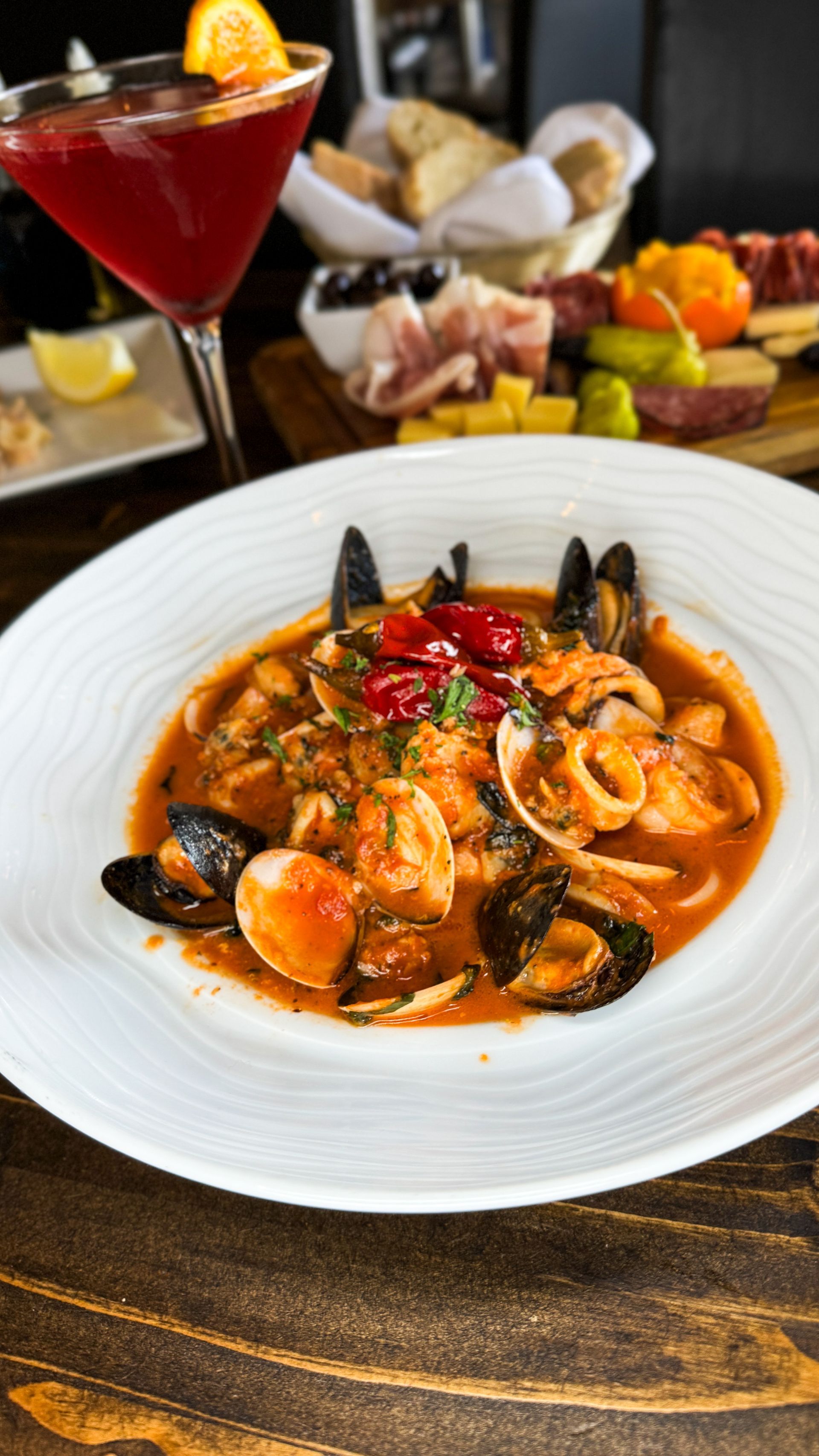 Seafood stew in white bowl, with mussels, calamari, and clams; a cocktail and charcuterie board in background.