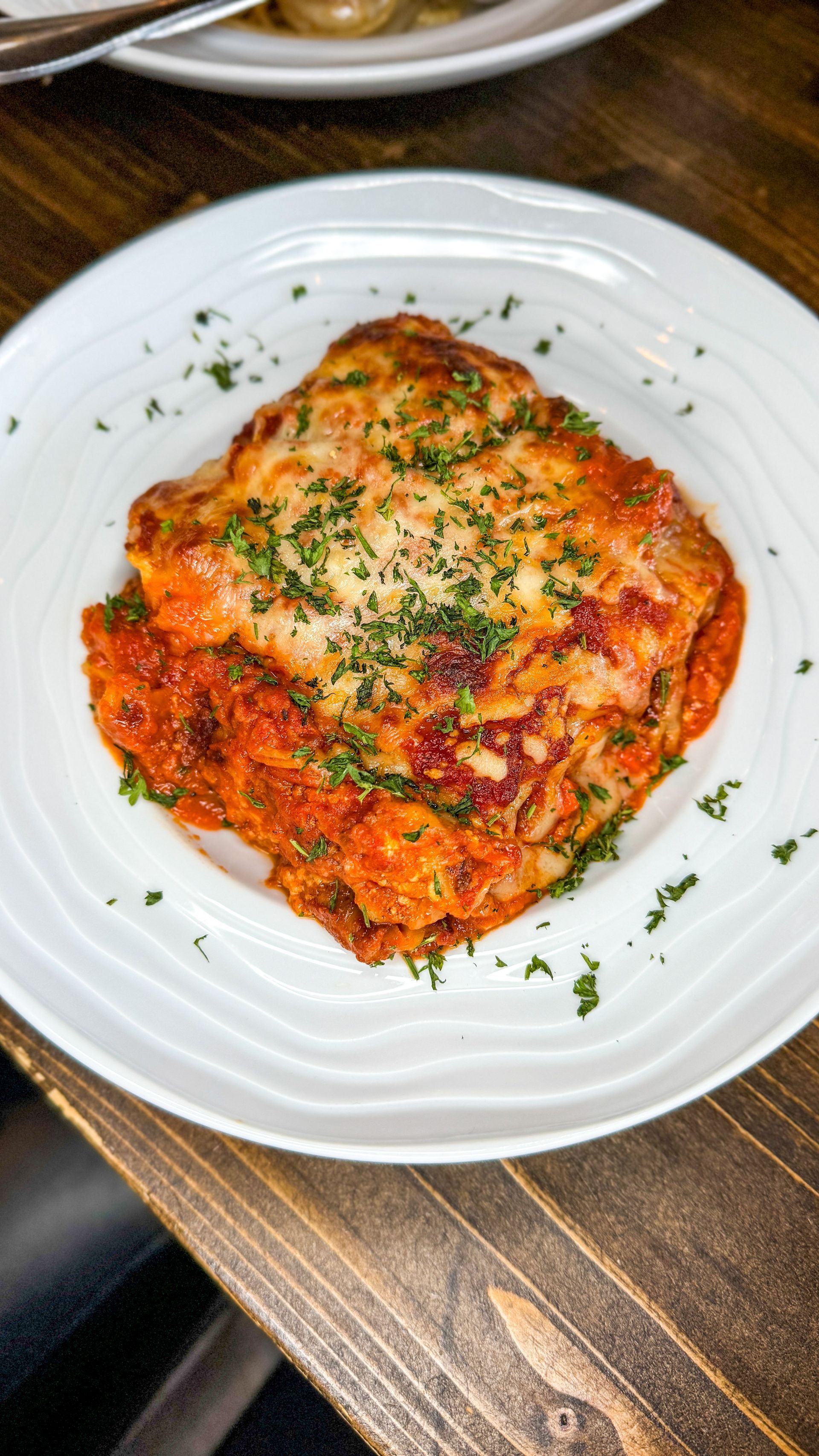 Lasagna in a white bowl, topped with melted cheese and parsley, on a dark wooden table.