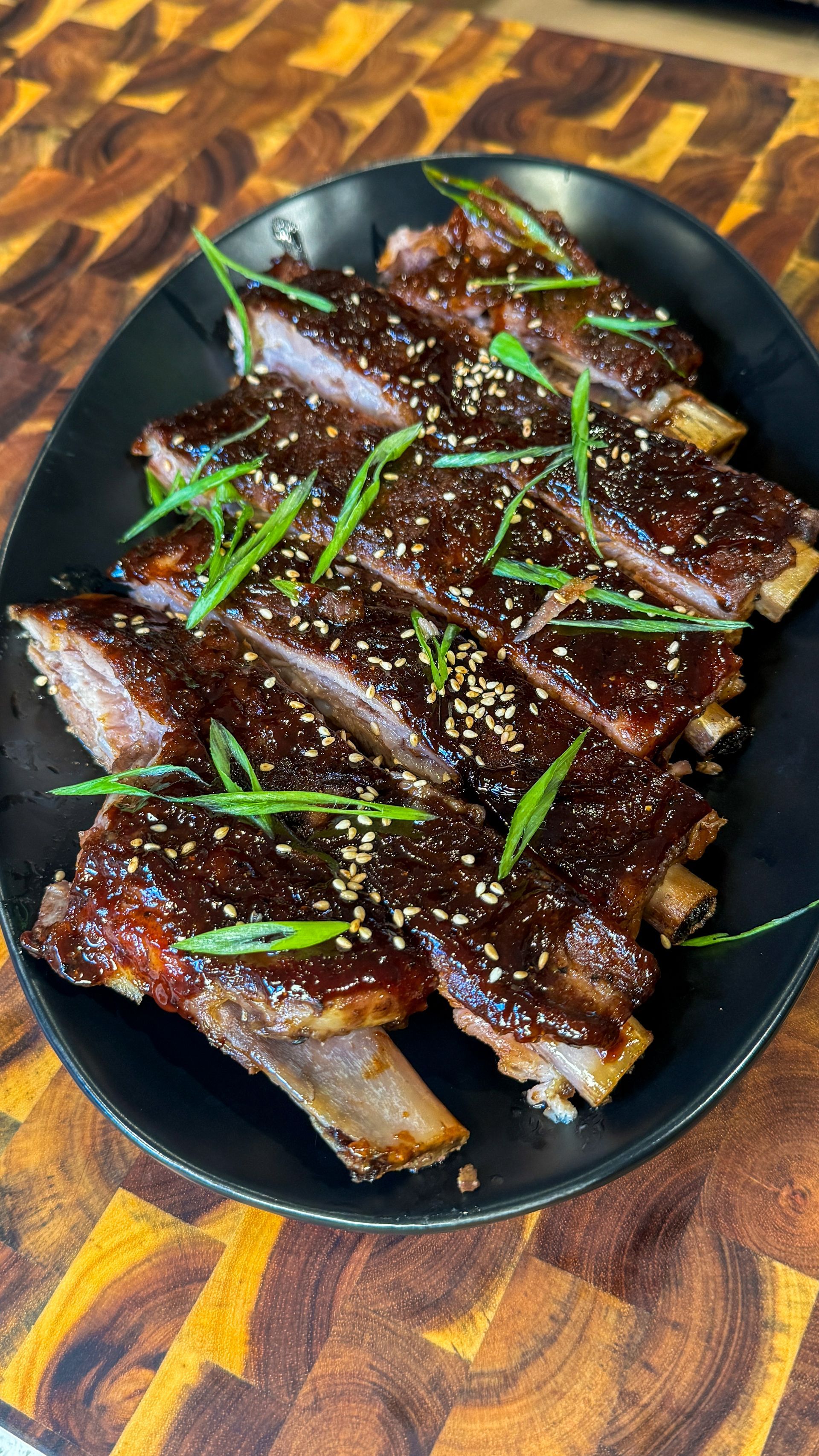 Glazed ribs on a black plate, garnished with green onions and sesame seeds, on a wooden surface.