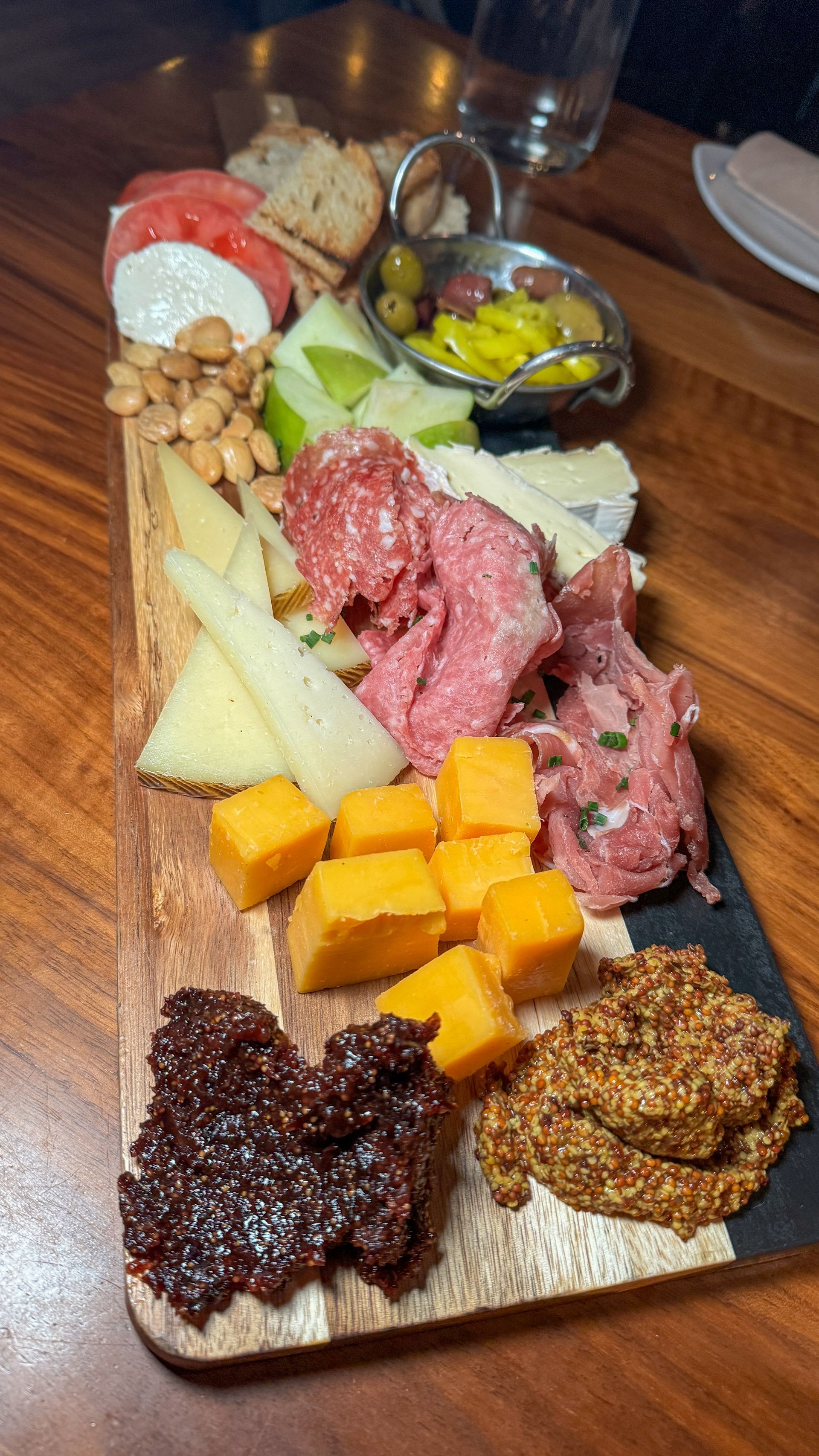 A wooden cutting board filled with meat , cheese , nuts , and olives on a wooden table.