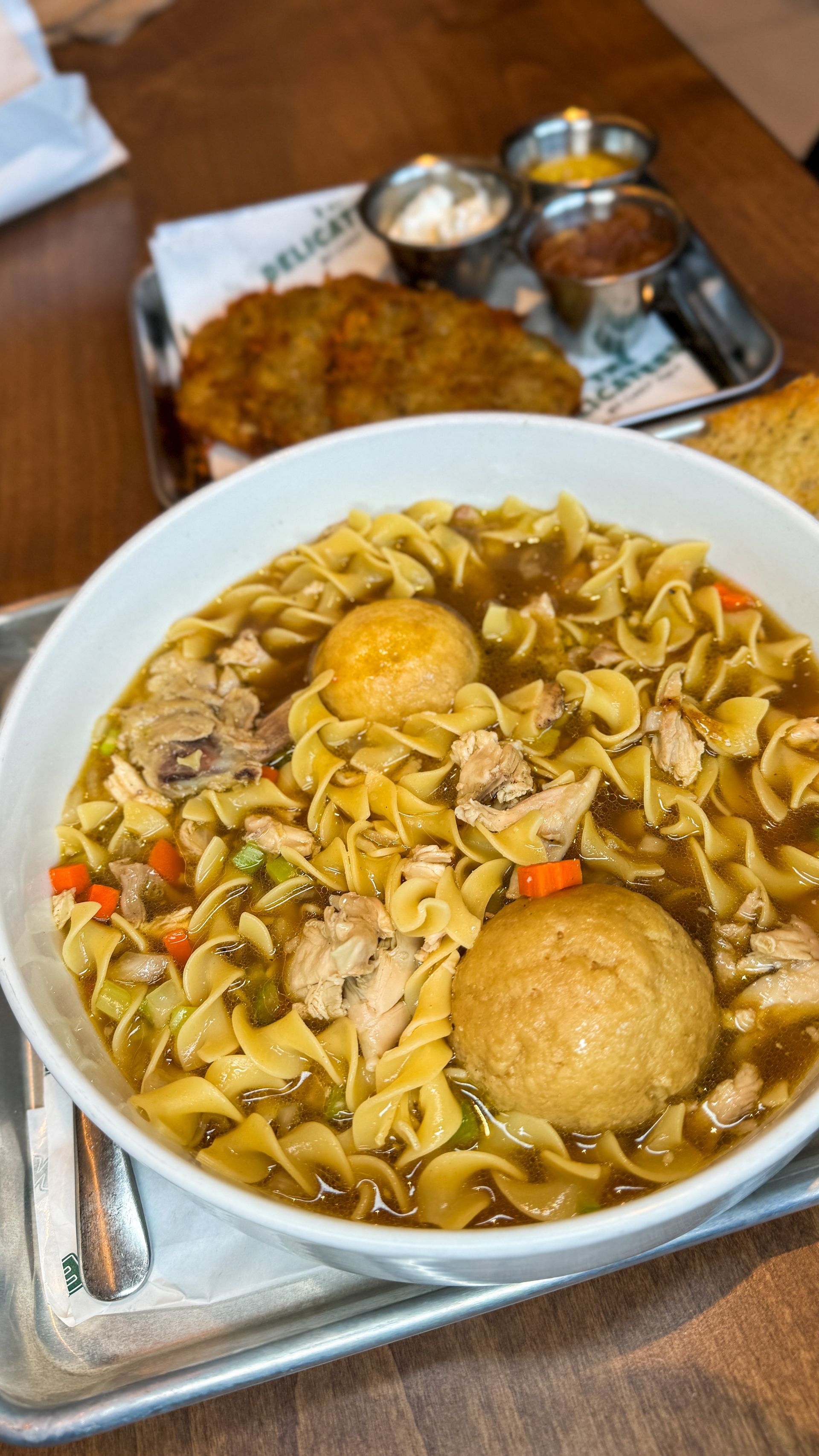 Bowl of chicken noodle soup with matzah balls, fried potato cake, and dipping sauces.